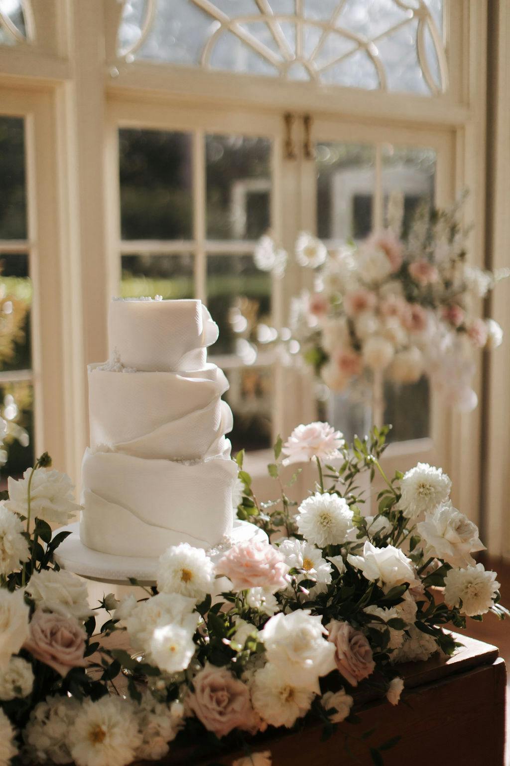 A white, three-tier wedding cake with a modern, asymmetrical design sits on a wooden table adorned with white and light pink flowers. Behind it, a large window with intricate panes allows soft sunlight to filter in, illuminating a floral arrangement in the background.