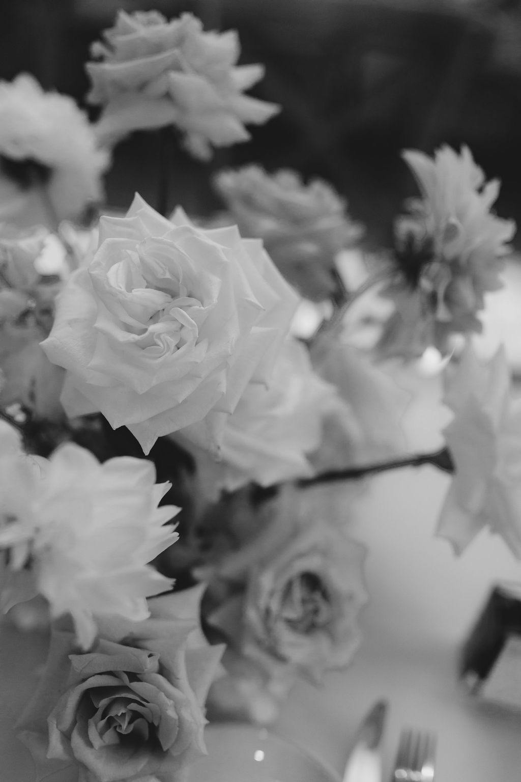 Black and white image of a bouquet of roses and other flowers on a table, with blurred background. The flowers are in various stages of bloom, creating a classic and elegant arrangement. Cutlery and part of a plate are visible in the foreground.