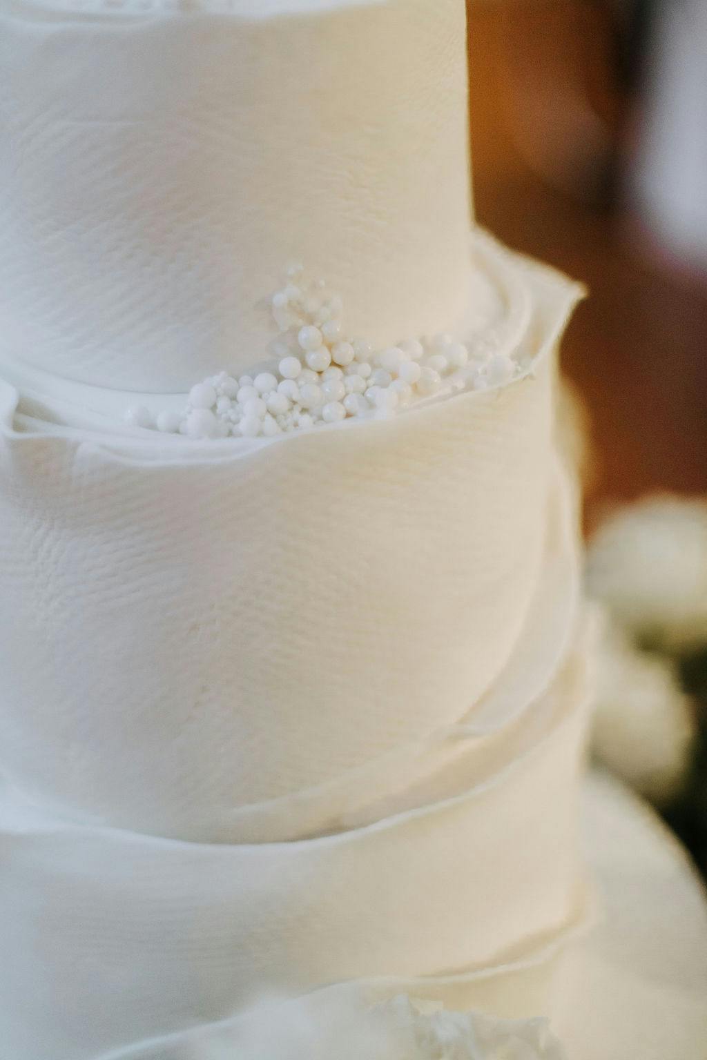 A close-up of a multi-tiered cake decorated to resemble rolls of toilet paper. The cake features realistic textures and detailing, with white fondant rolled and layered to mimic the appearance of toilet paper. Small white decorative elements add a touch of elegance.