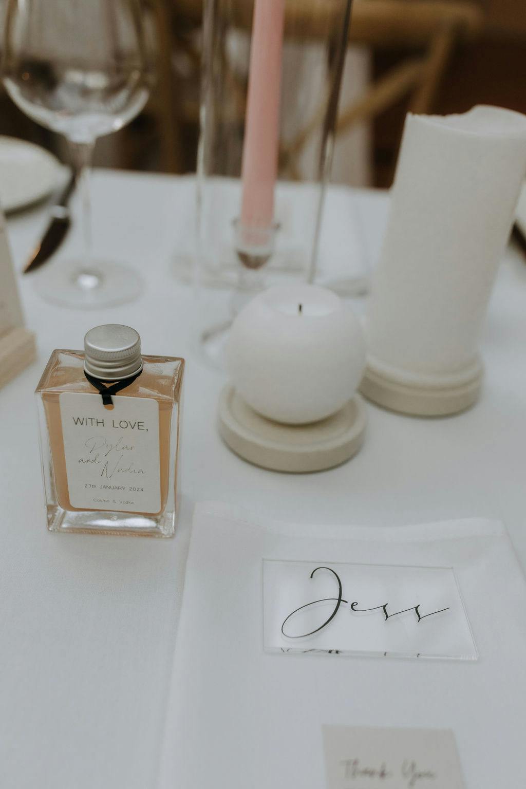 Close-up of a wedding table setting featuring a small bottle labeled "With Love..." a candle, a tall pink taper candle, glassware, and a napkin with a name card that reads "Jess." The setup is elegant with neutral tones and a minimalist aesthetic.