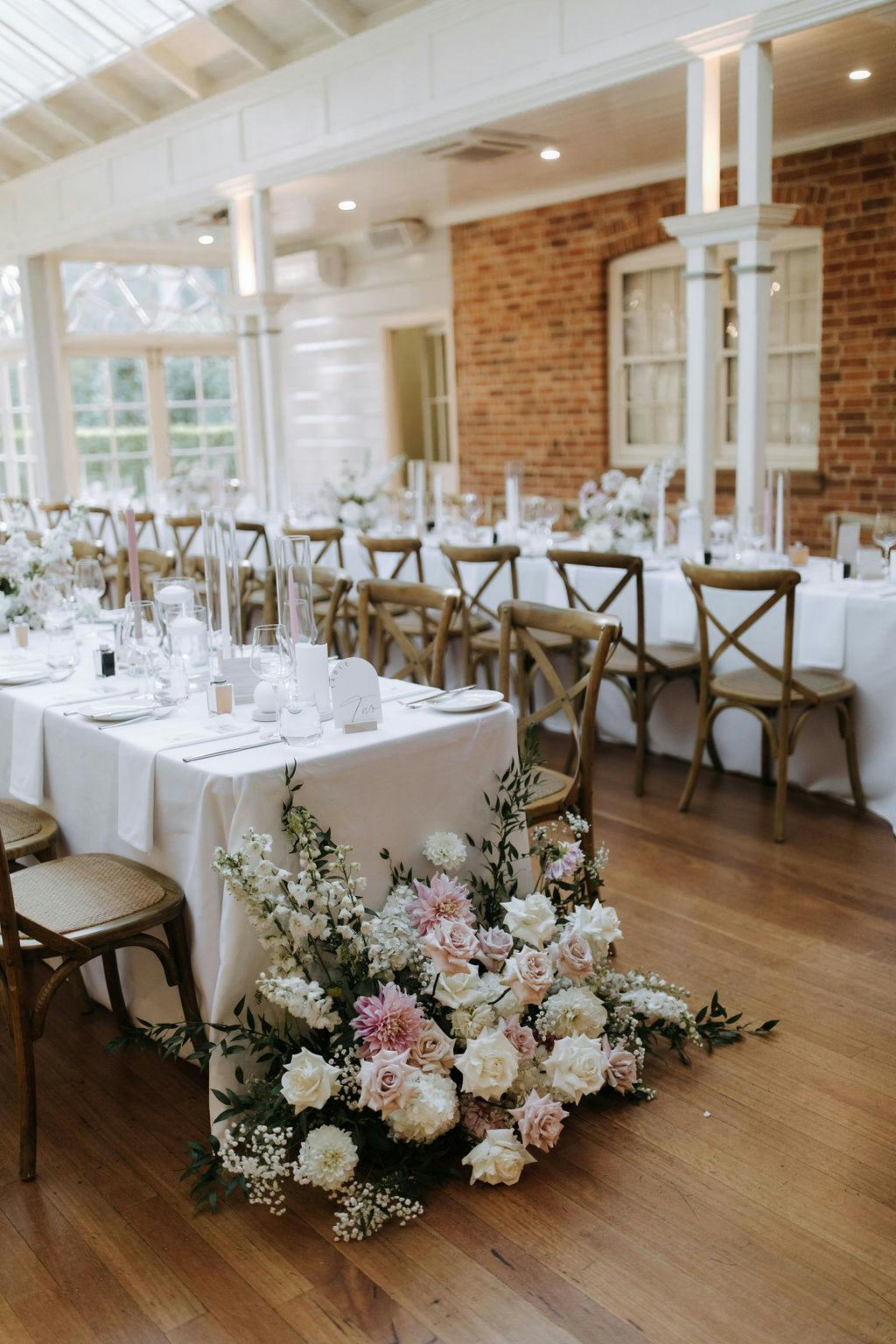 A beautifully decorated wedding reception room features a long table with white linens, set with glassware, white plates, and tall clear vases with candles. A floral arrangement of pink and white flowers with greenery sits at the end of the table.