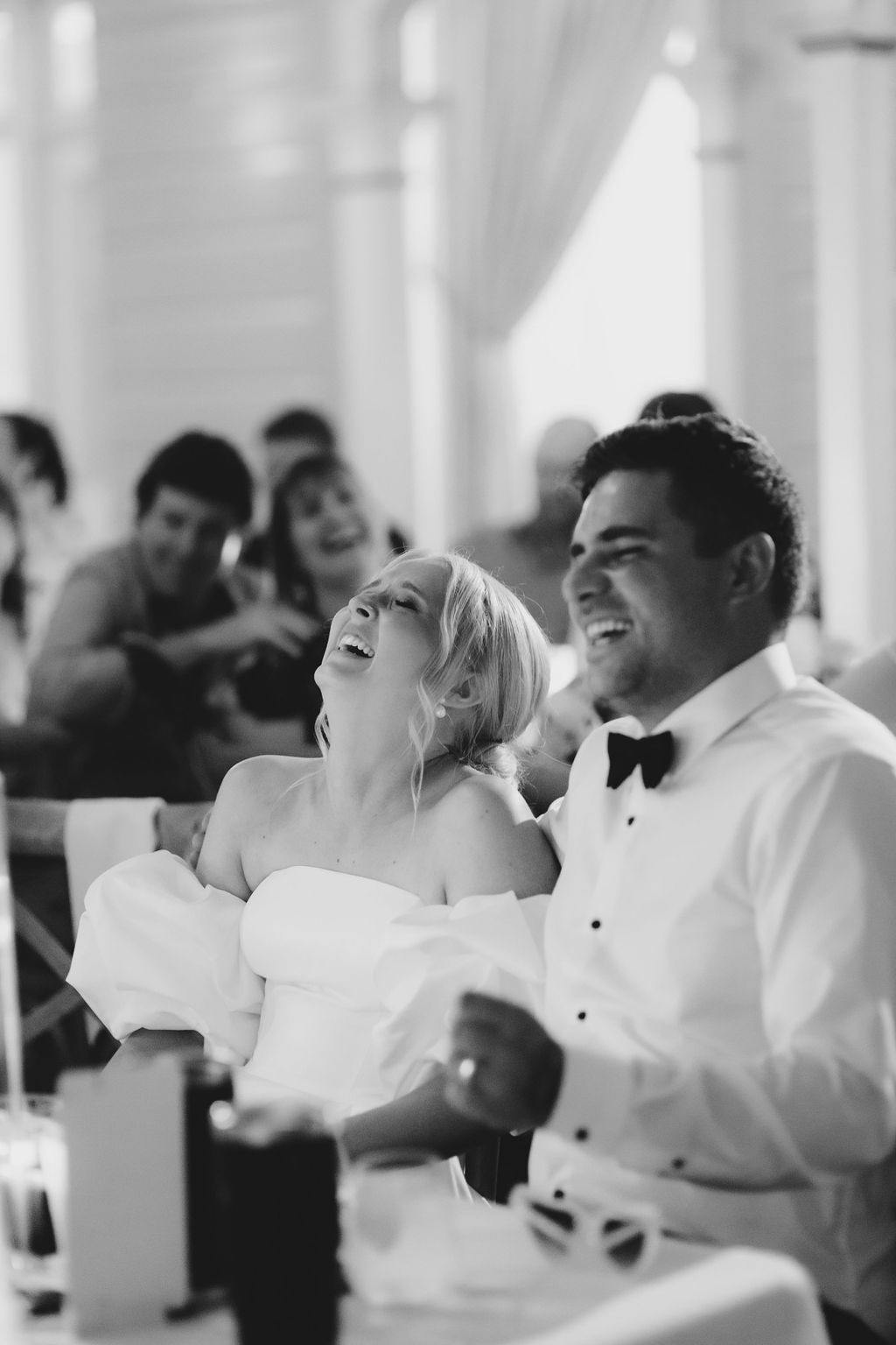 A joyful bride and groom, both dressed in formal attire, sit at a reception table, laughing heartily. The bride is in an off-shoulder white dress and the groom in a white shirt with a black bow tie. Guests in the background also appear to be smiling and enjoying the moment.