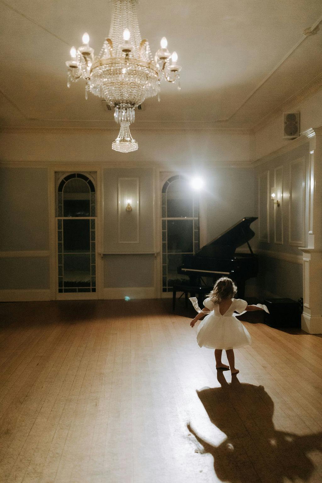 A young child in a white dress dances alone in a dimly lit, elegant room with high ceilings, a polished wooden floor, and classic wall paneling. A grand piano and an ornate chandelier add to the room's grandeur, while the child's shadow is cast on the floor.