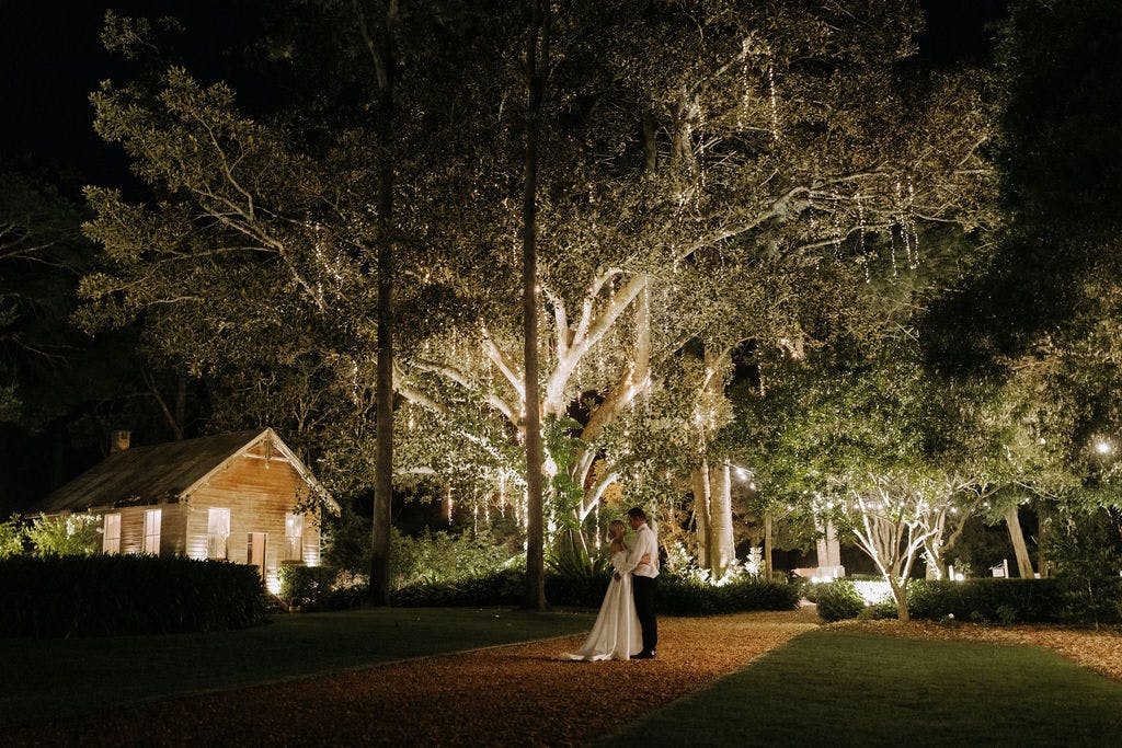 newlyweds posing for a photograph in the grounds of gabbinbar at night, the trees are glowing from all the lights hanging as decoration