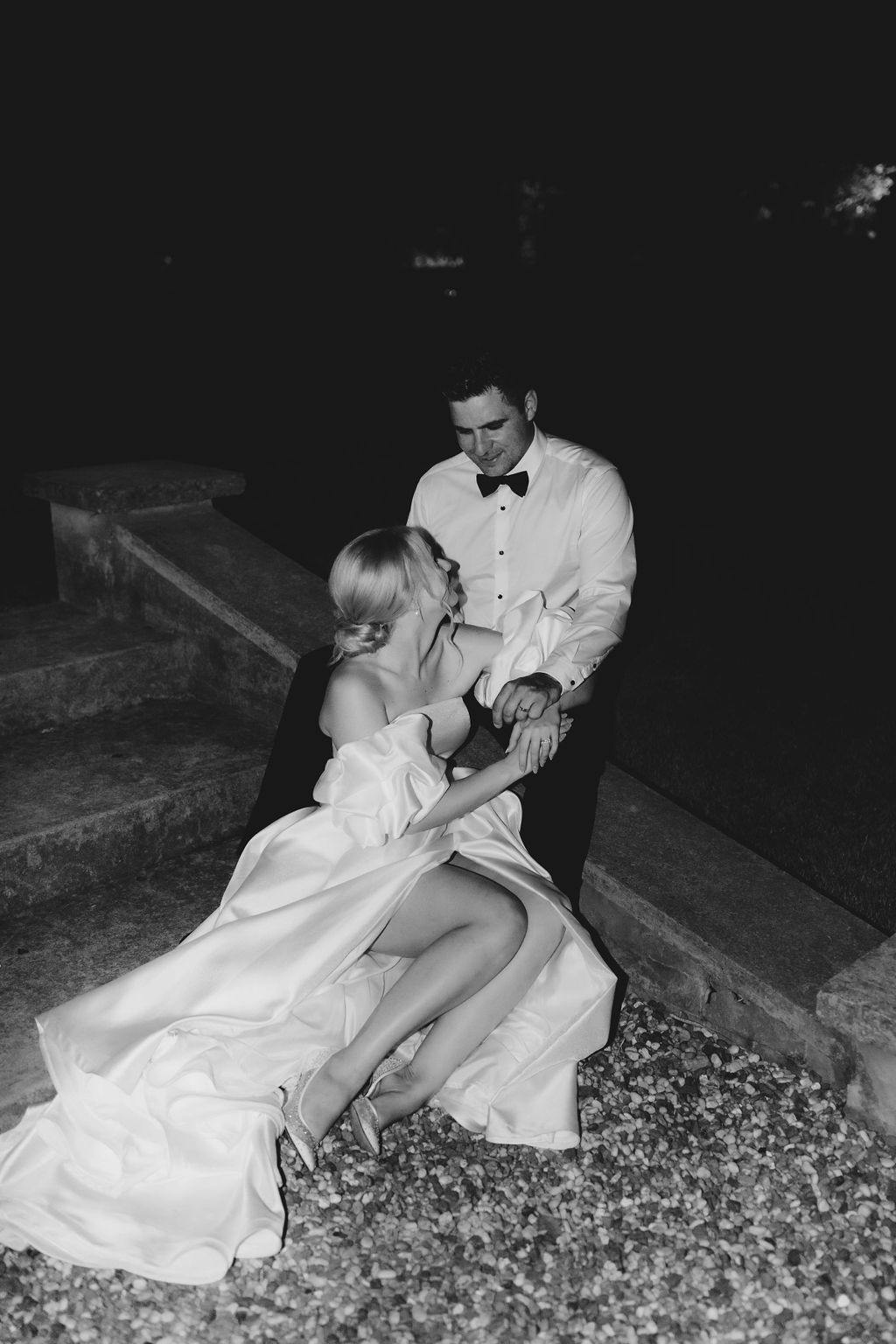 A black-and-white photo of a couple sitting on stone steps at night. The woman is wearing a white, off-shoulder dress with a flowing train, and the man is in a white shirt with a black bow tie. They are holding hands, sharing an intimate moment.