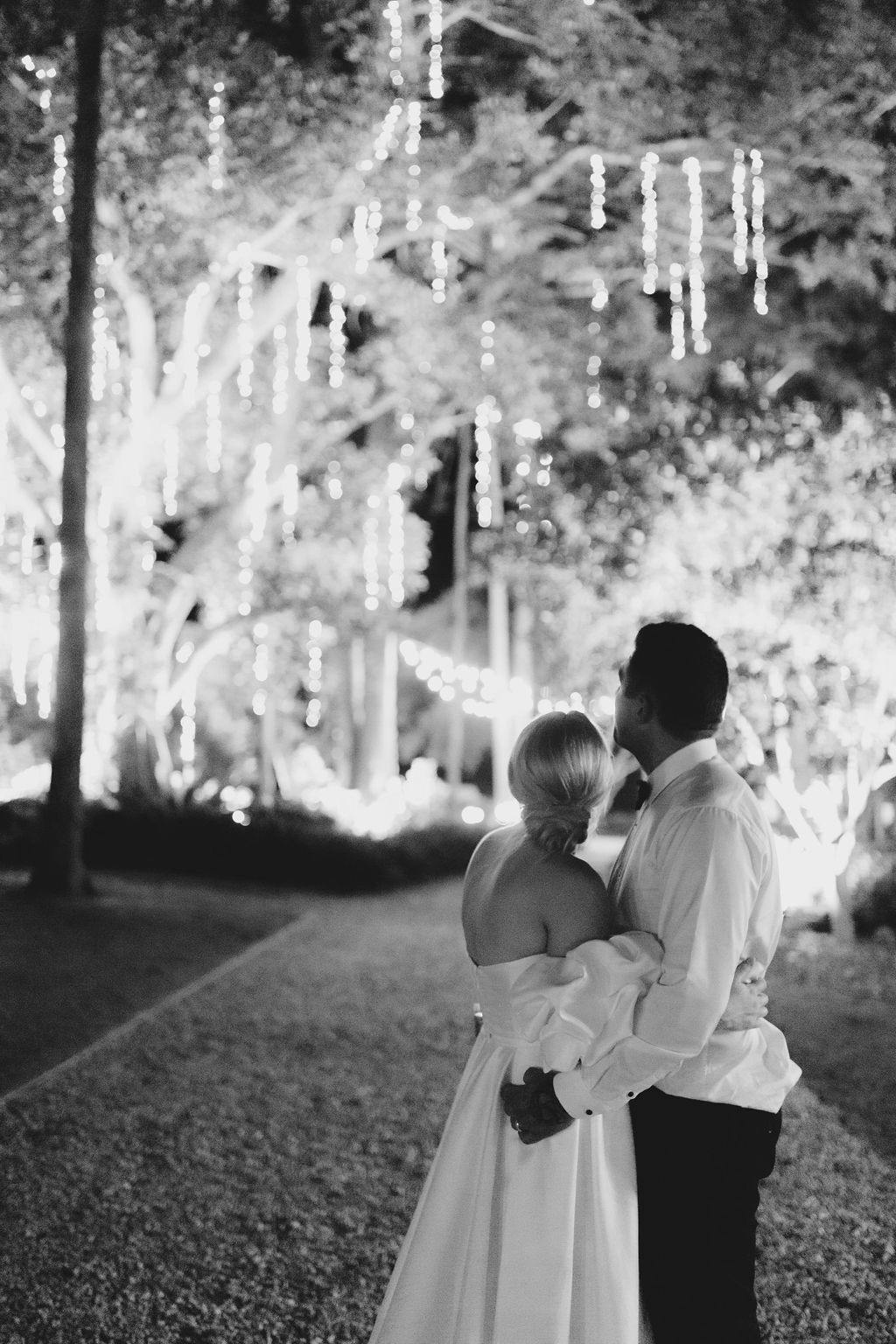 A black-and-white photo of a couple embracing from behind. They are facing a pathway adorned with hanging string lights draped over trees. The woman is wearing a strapless gown, and the man is dressed in a shirt and pants. The scene is calm and romantic.