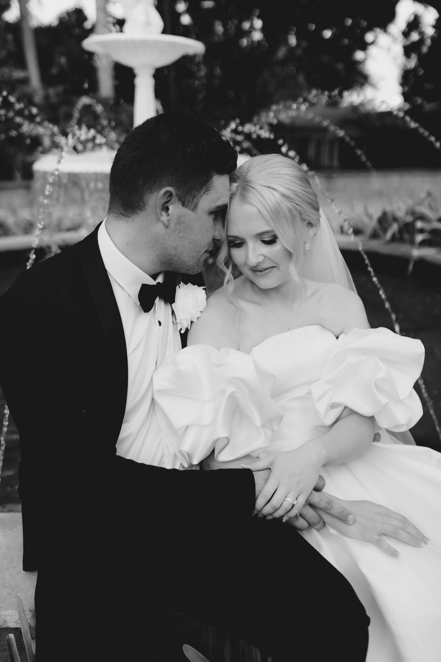 Black and white photo of a newlywed couple seated closely on a bench. The groom, in a suit, leans his forehead against the bride's head. The bride, in an off-shoulder wedding dress, smiles gently. A water fountain is visible in the background.