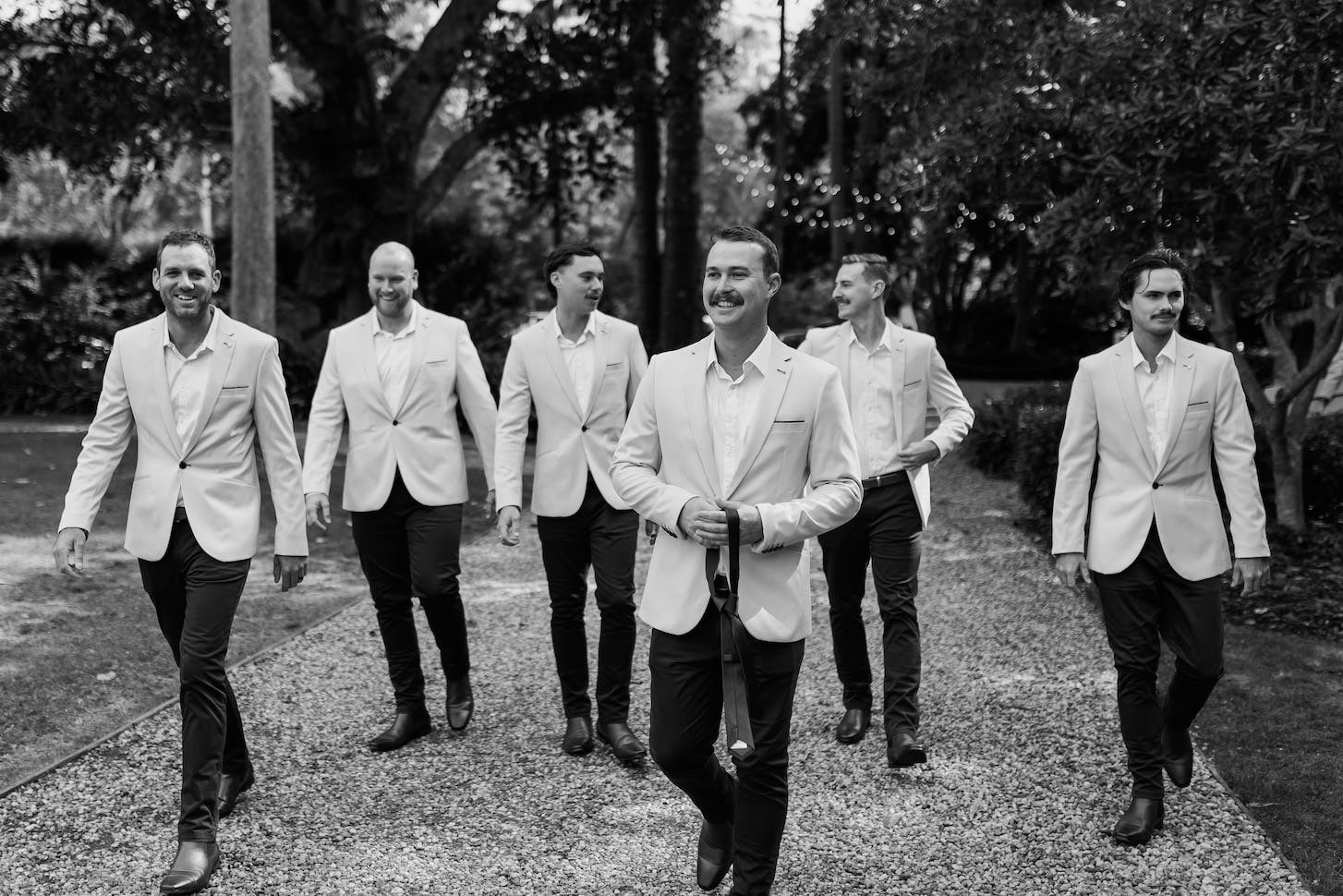 A black and white photo of six men walking on a gravel path outdoors, surrounded by trees. They are dressed in light-colored blazers and dark pants, with the man in the center holding a champagne bottle. The group appears to be smiling and enjoying the moment.