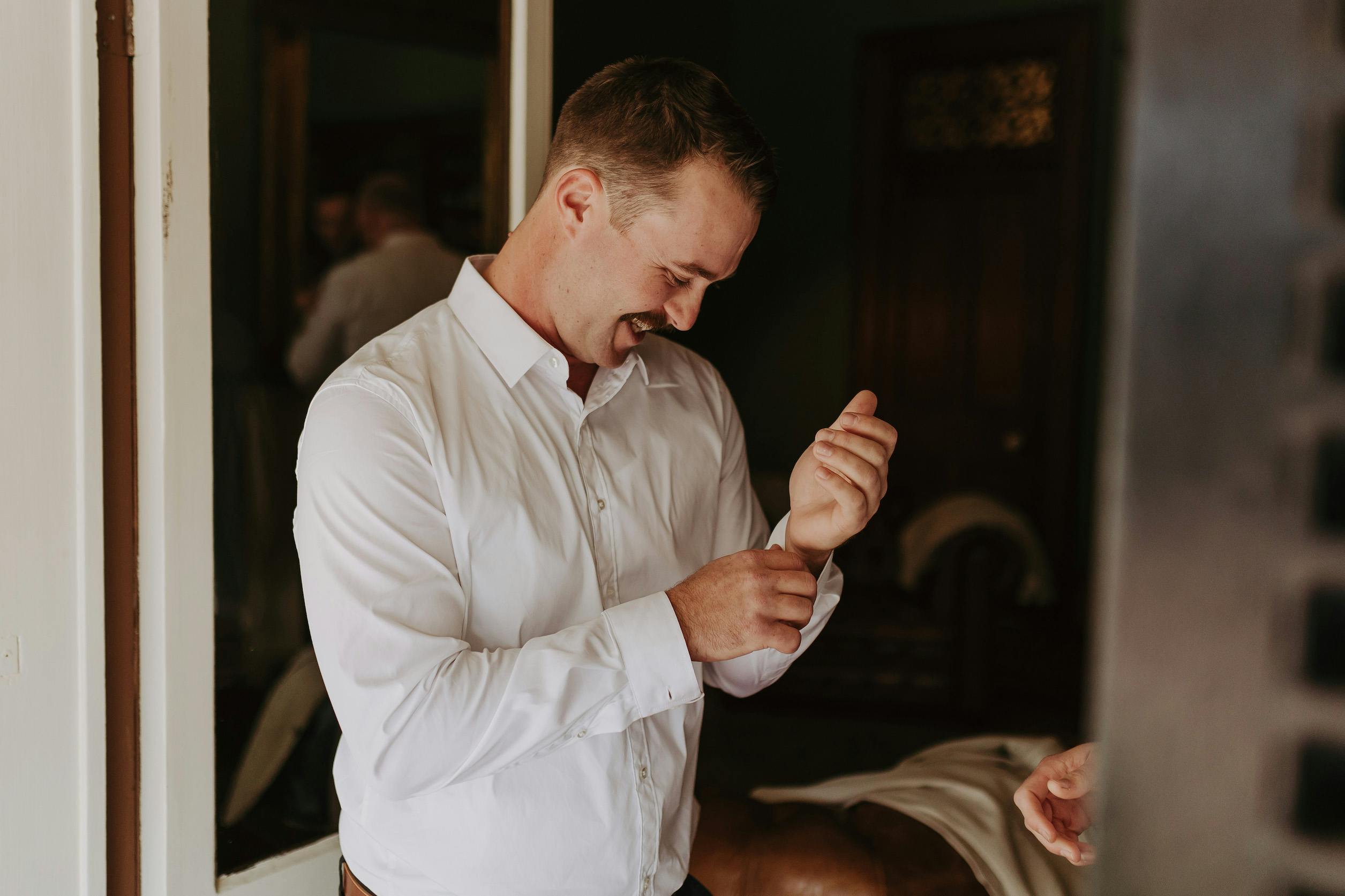 A man stands in a room adjusting the cuff of his white shirt with a slight smile. He has short hair and is dressed neatly. The room's background includes a mirror and a wooden door, with soft indoor lighting enhancing the warm atmosphere.