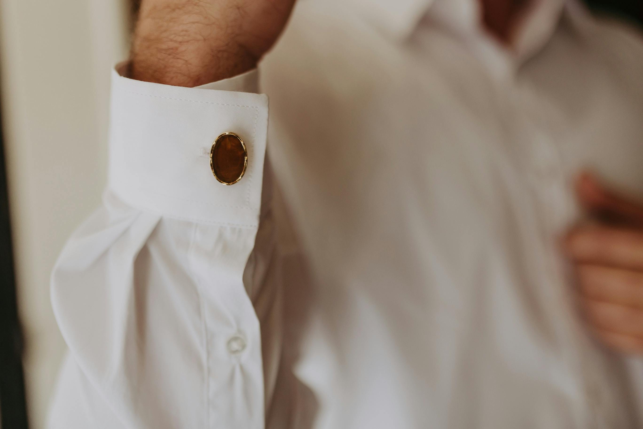 A person wearing a white dress shirt with a gold cufflink on the cuff. The image is closely cropped to focus on the cufflink and part of the arm. The background is blurred, making the cufflink the central element of the photo.