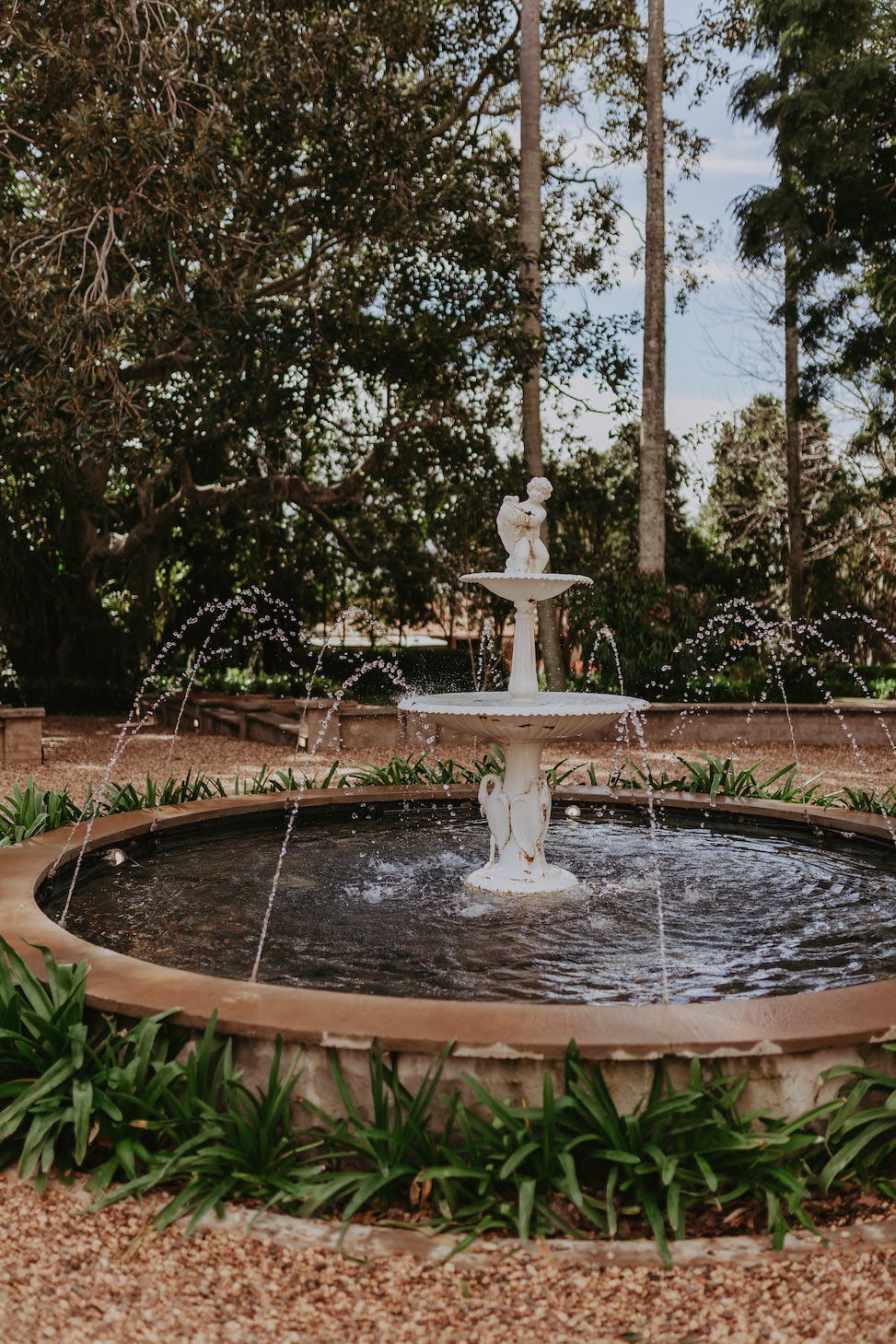 A white, tiered stone fountain with water jets sprays in a circular basin. Surrounded by lush greenery and flowers, the fountain is set against a backdrop of tall trees and a bright sky. The scene is tranquil and natural, with pathways around the fountain.