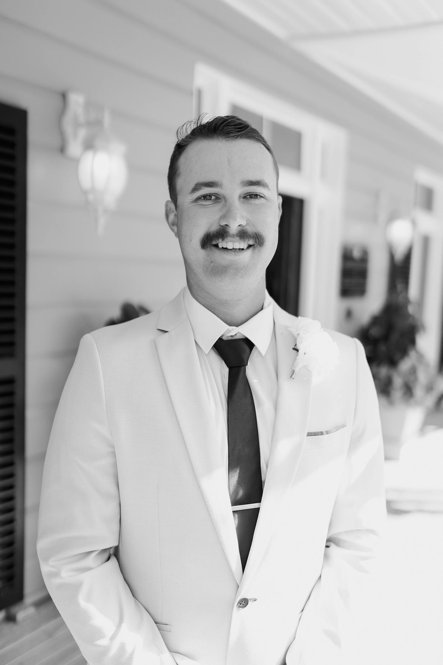 A man dressed in a light-colored suit with a dark tie and a boutonniere smiles at the camera. He has a mustache and is standing outside a building with siding and windows. The photo is in black and white.