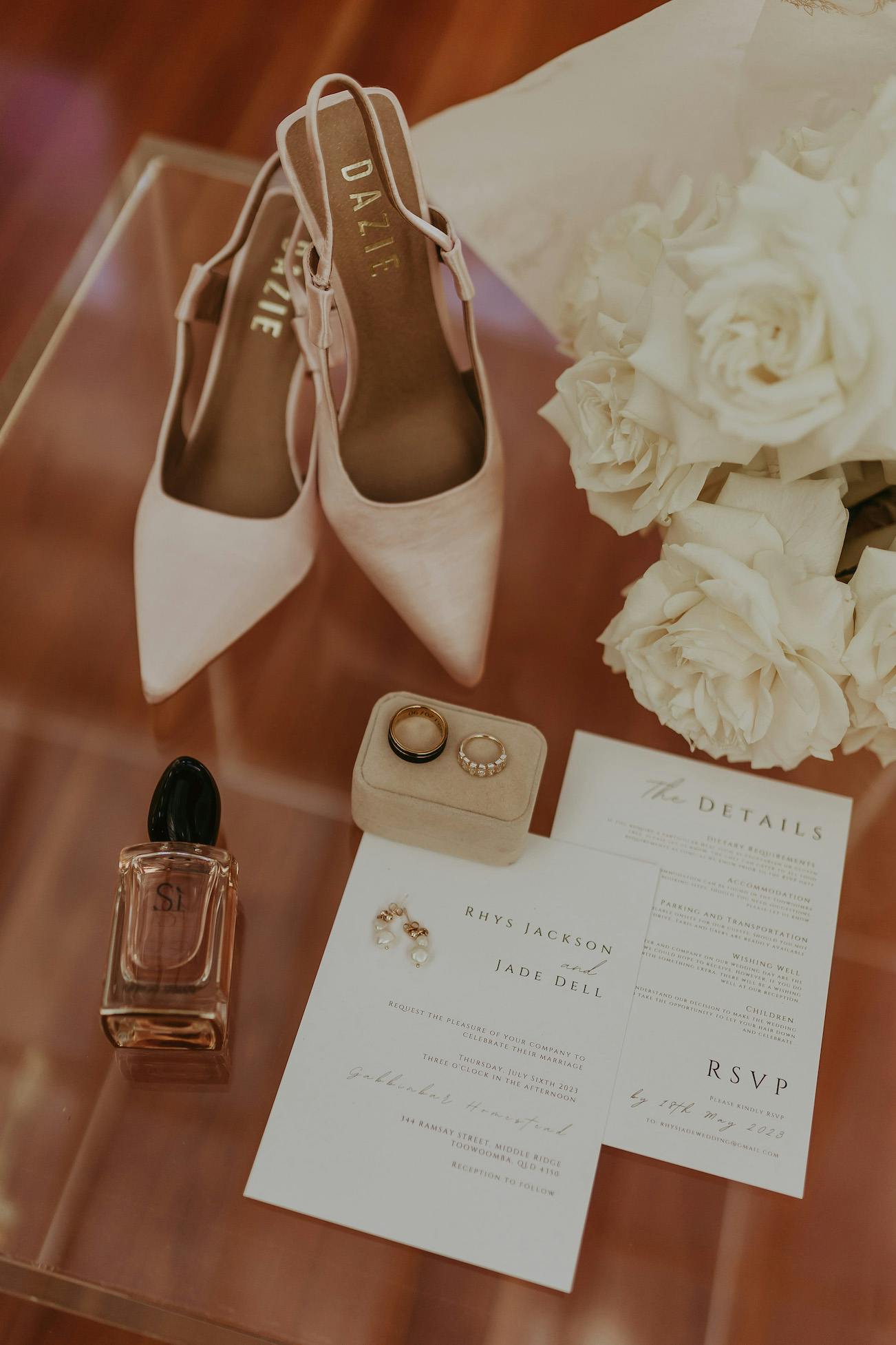 A wedding flat lay with blush pink high heels, gold rings, pearl earrings, a perfume bottle, and white roses. Also included are wedding details cards with RSVP information, featuring the names Rhys Jackson and Jade Dill.