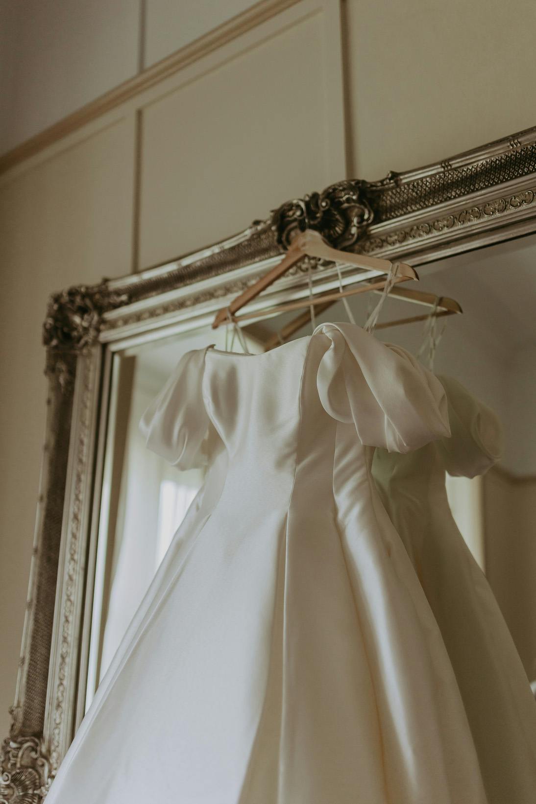 A white satin wedding dress with short puffy sleeves hangs on a wooden hanger in front of an ornate, large vintage mirror with a detailed silver frame. The dress reflects softly in the mirror, creating a romantic and elegant atmosphere.