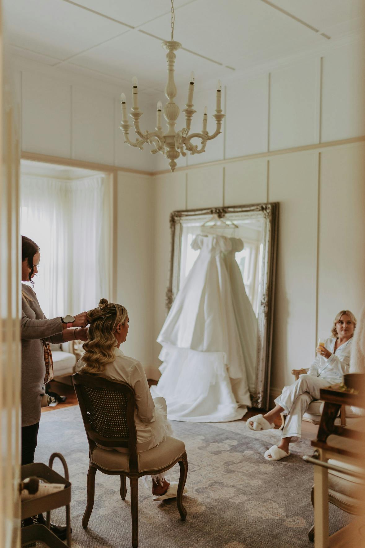 A bride sits on a chair while getting her hair styled by another person, with a long white wedding dress hanging on a large ornate mirror in the background. Two other women dressed in white robes are sitting on a couch in the cozy, elegantly decorated room.