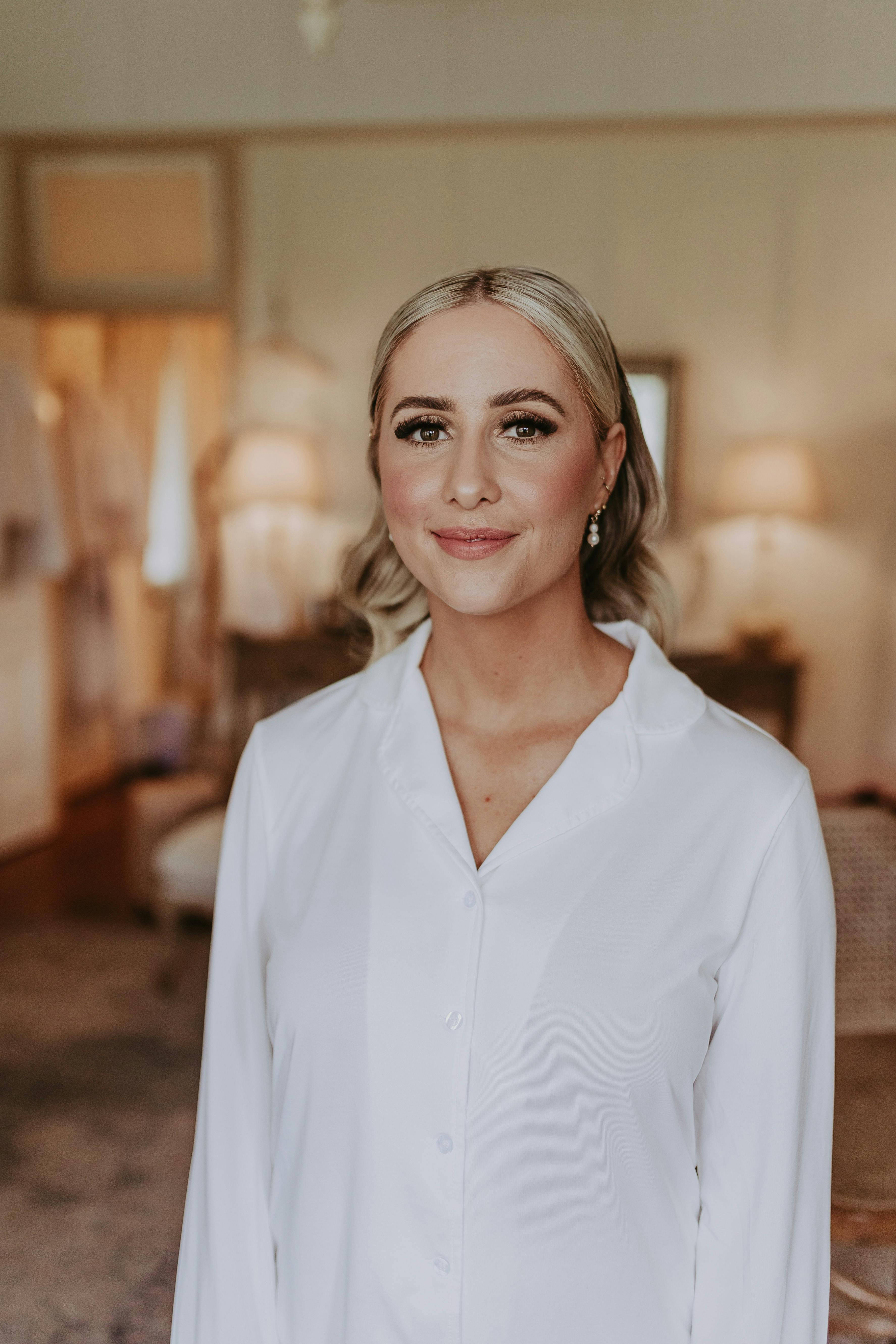 A woman with blonde hair styled in loose waves stands indoors wearing a white button-up shirt. She has a warm smile and is in a softly lit room with furniture and décor in the background.