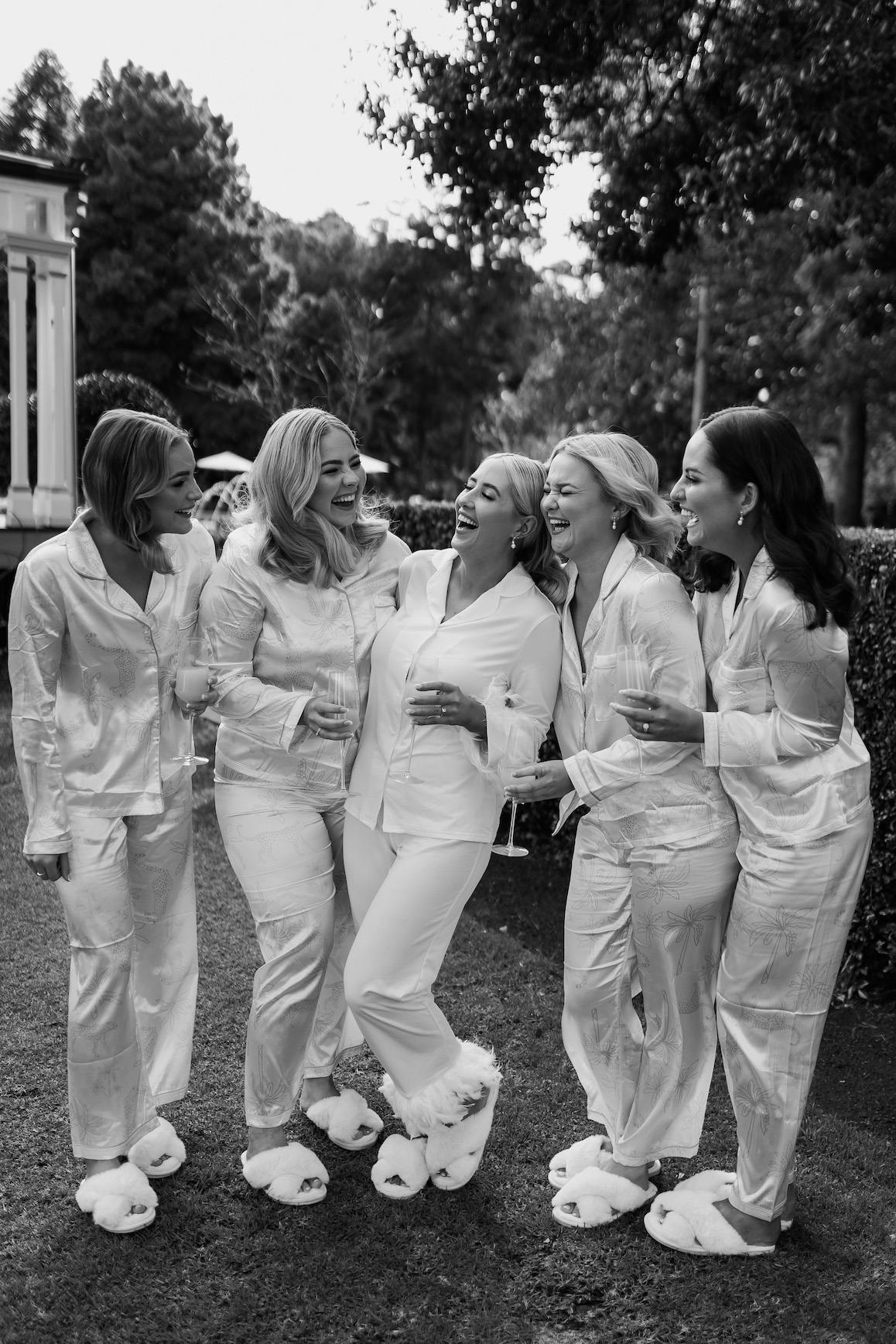 A group of five women dressed in matching white pajamas and fluffy slippers stand outdoors, joyfully laughing and talking. Some are holding champagne glasses. The background features trees and part of a building. The scene is in black and white.