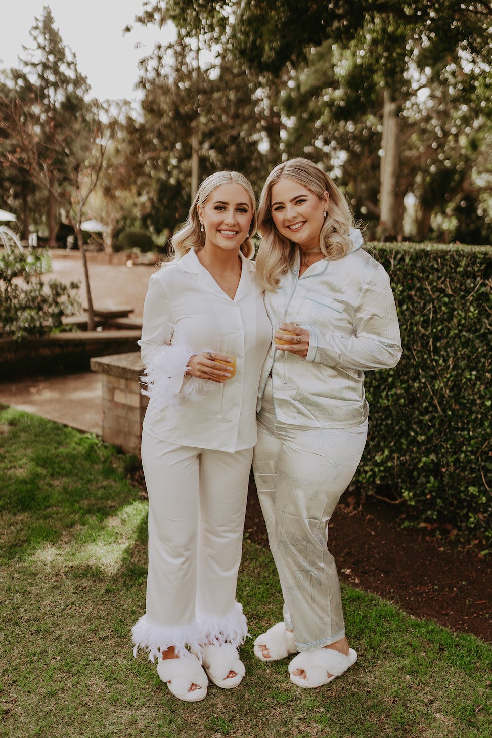 Two smiling women stand outside on grass, wearing coordinated white pajama sets. One has feathered cuffs on her pants and the other is in satin pajamas. They each hold a drink in one hand and appear relaxed. Greenery and tree-lined background is visible.
