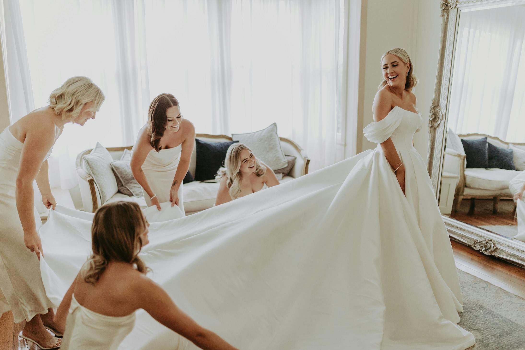 A bride in an off-the-shoulder wedding dress stands smiling while four bridesmaids in matching white dresses gather around her, helping with her long train. They are in a bright room with a large window and a full-length mirror.