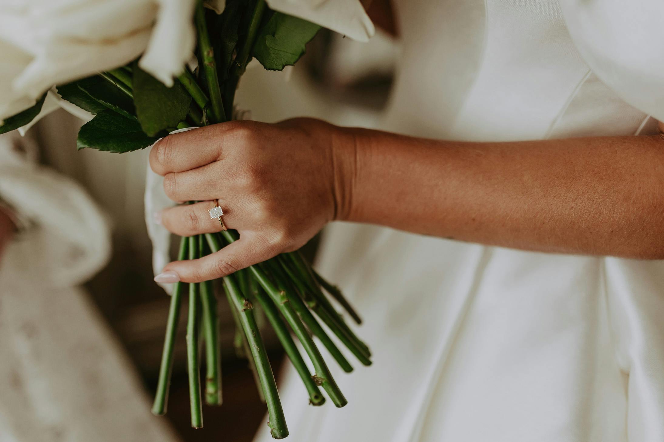 A close-up of a person’s hand holding a bouquet of white flowers. The person is wearing a white outfit, and visible on their ring finger is a diamond engagement ring. The background is blurred, keeping the focus on the hand, flowers, and ring.
