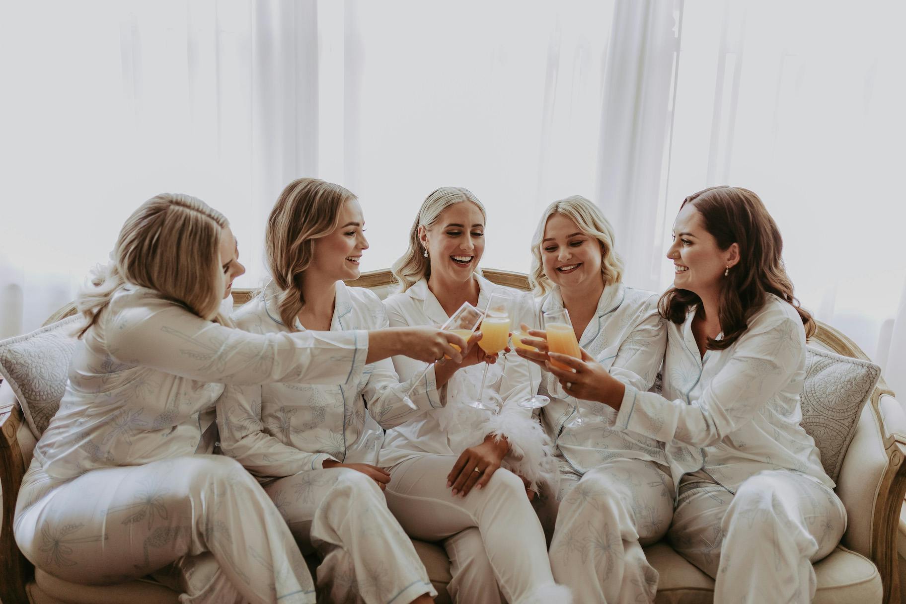 Five people in matching white pajamas are sitting on a couch, smiling and clinking glasses of orange juice in a cheerful toast. They appear to be celebrating a special occasion, like a wedding morning or a bachelorette party, in a bright room with light curtains.