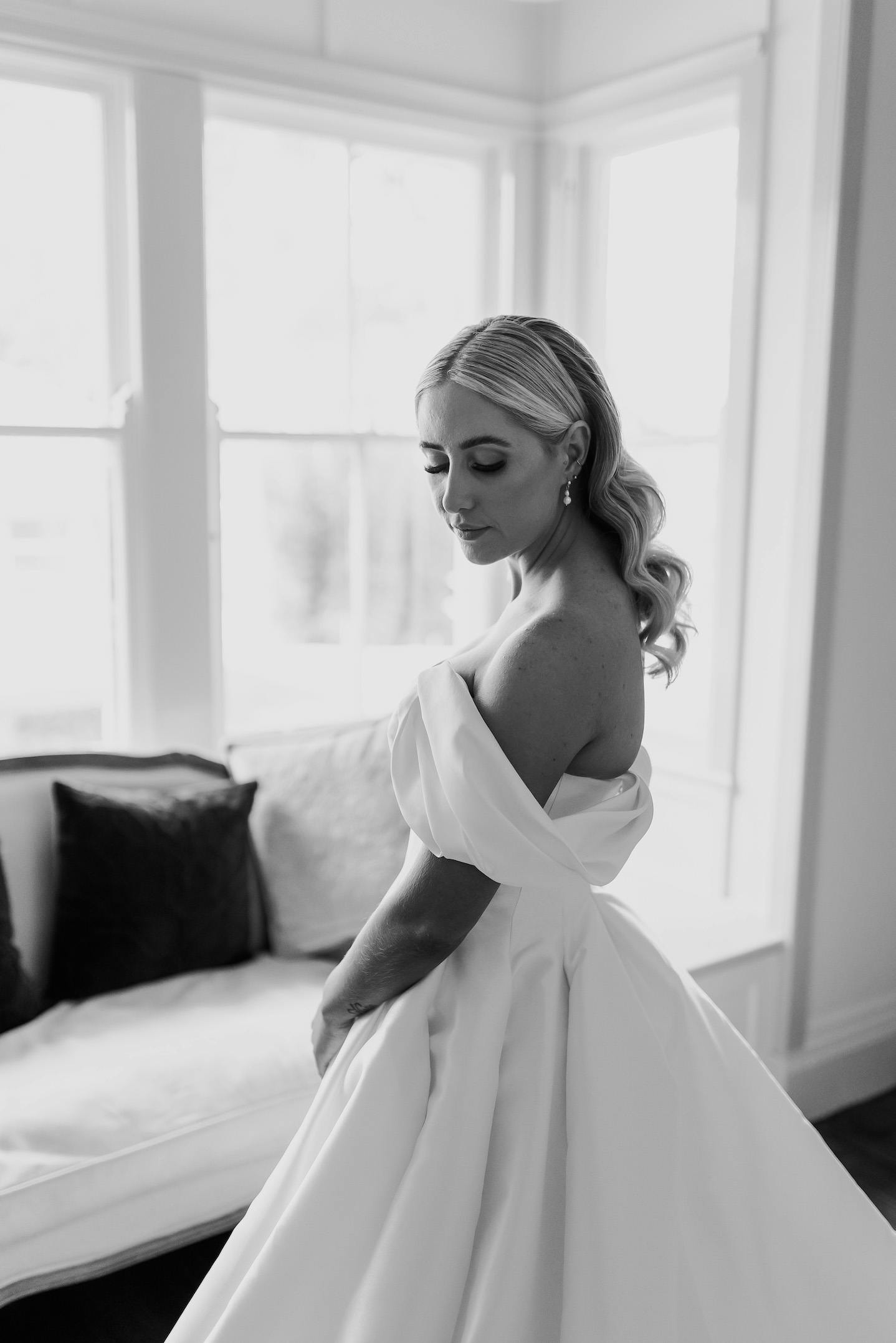 Black and white photo of a bride in an off-the-shoulder wedding gown, standing in front of a window. She has long, wavy hair and is looking down with a serene expression. Behind her, there is a cushioned bench with pillows.