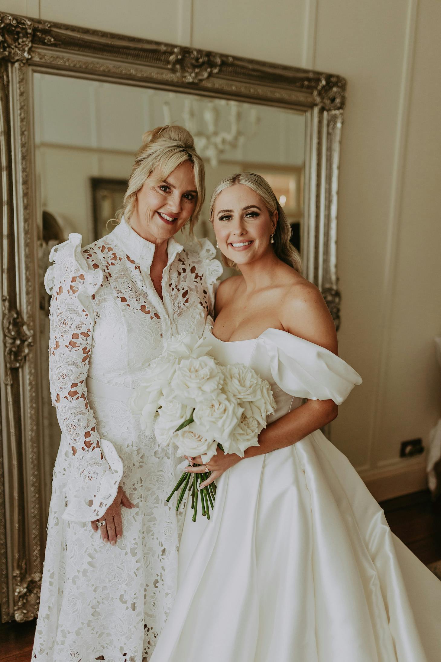 A bride and an older woman smile together in front of an ornate mirror, dressed in white. The bride wears an off-the-shoulder gown and holds a bouquet of white roses. The older woman is in a lace dress with ruffled sleeves. Both are standing in an elegant room.