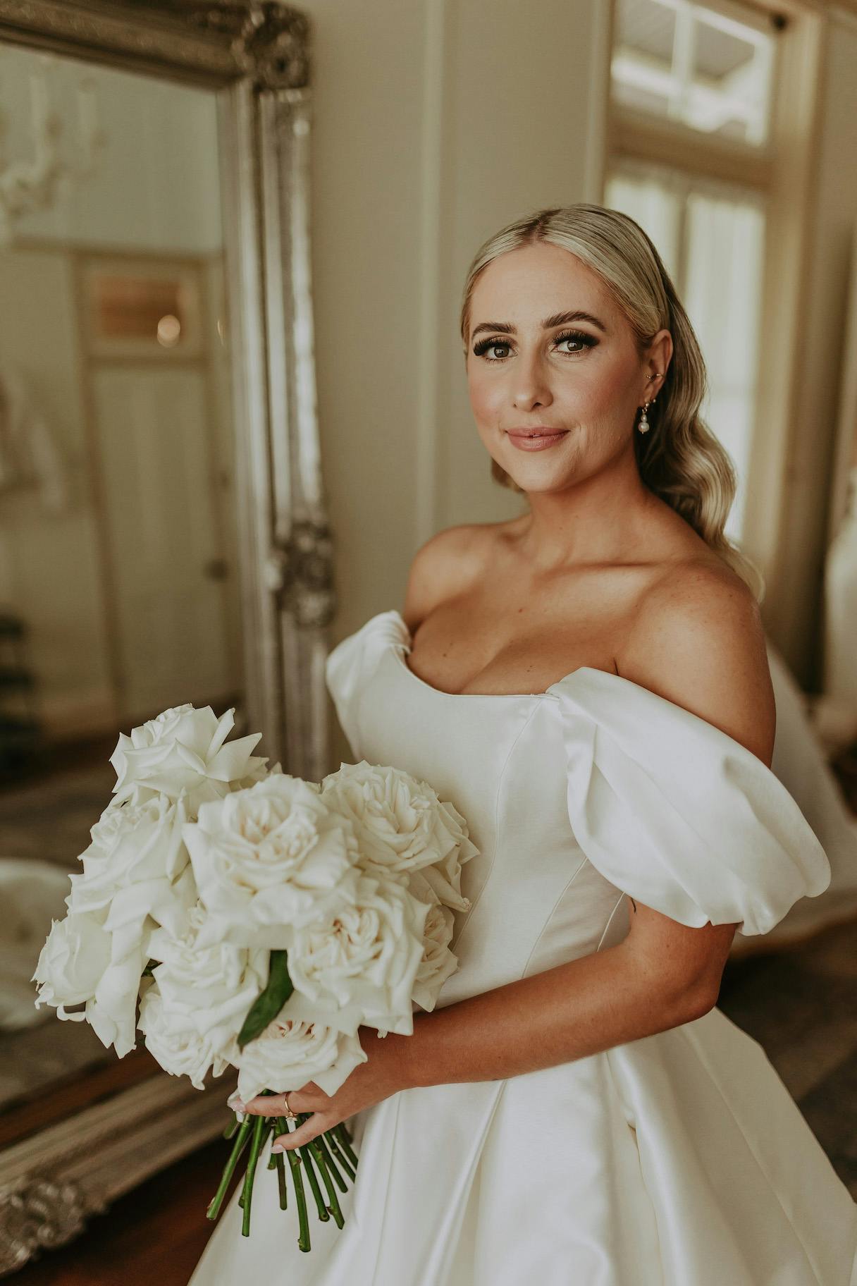 A bride wearing an off-the-shoulder white wedding dress holds a bouquet of white roses. She stands in front of a large ornate mirror and smiles softly, with blonde hair styled in loose waves. The room behind her has elegant decor and soft lighting.