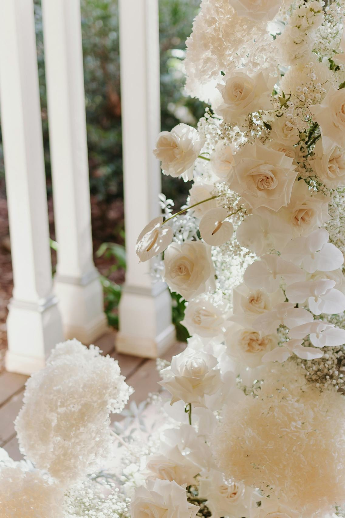 A close-up of a floral arrangement featuring a variety of white flowers, including roses and baby's breath. The arrangement is set against the backdrop of a white porch railing with greenery in the background. The overall appearance is elegant and serene.