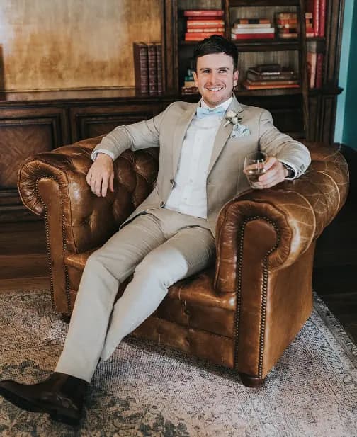 A man in a beige suit with a blue bow tie sits in a brown leather armchair, holding a glass of whiskey and smiling. Bookshelves and a wooden wall adorn the cozy room's background.
