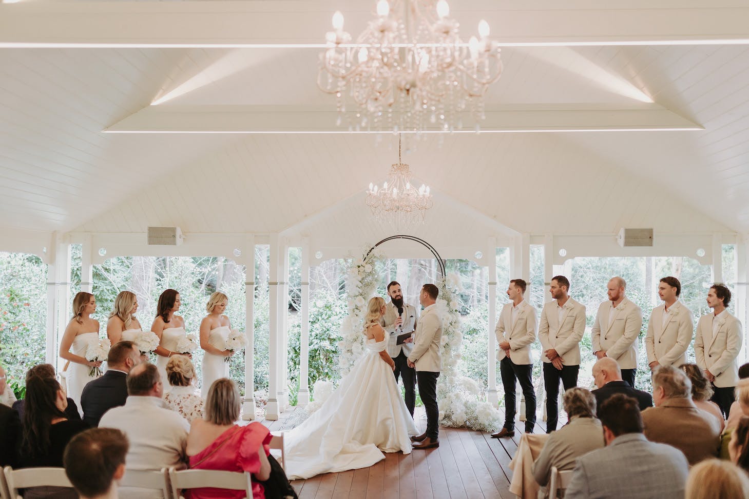 A wedding ceremony takes place in a charming, well-lit venue. The bride and groom stand at the altar with their wedding party lined up on either side. Guests are seated and watching the ceremony. Chandeliers hang from the ceiling, adding elegance to the setting.