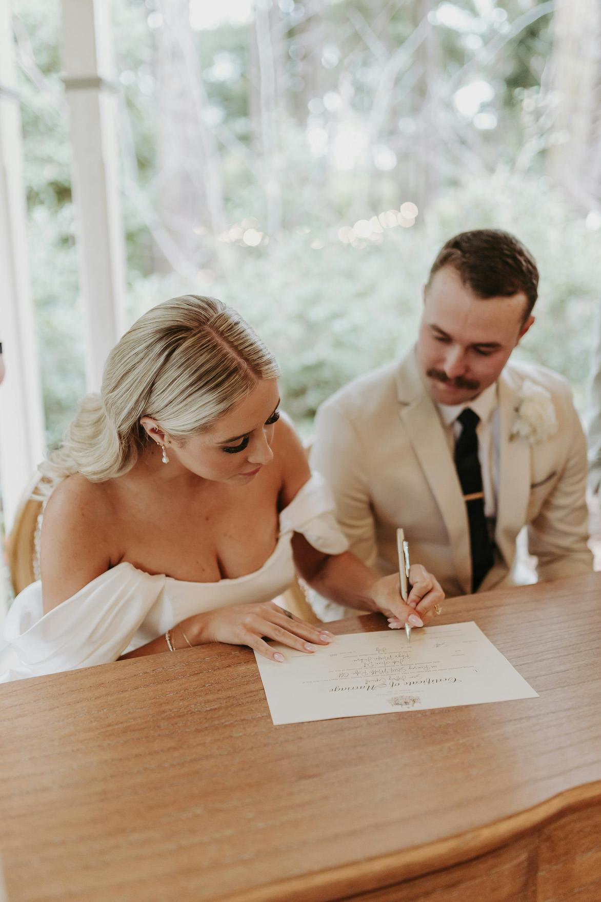 A bride and groom sit side-by-side at a wooden table. The bride, in an off-shoulder white dress, is focused on signing a document, while the groom, dressed in a beige suit and black tie, watches attentively. The background features greenery and a white structure.