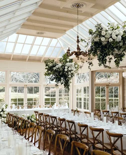A beautifully decorated indoor venue with a long dining table set for an event. The room features large windows and a glass ceiling, allowing natural light to flood in. Floral arrangements, featuring white flowers and greenery, are suspended from a vintage chandelier.