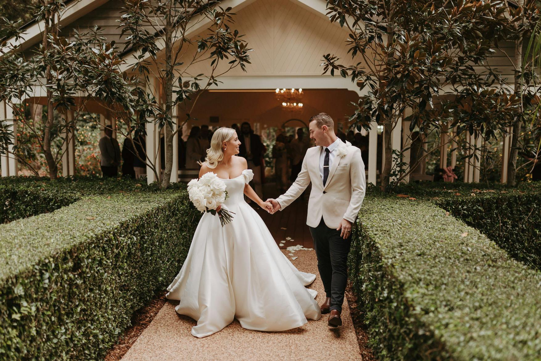 A newlywed couple walks hand in hand down a path lined with neatly trimmed hedges. The bride wears an off-the-shoulder white gown and holds a bouquet of white flowers, while the groom wears a light-colored suit with a tie. They are in front of a well-lit chapel.
