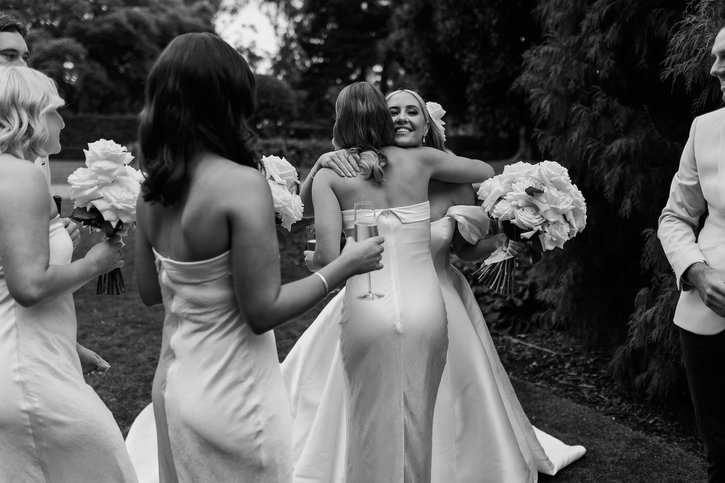 A bride in a white wedding dress warmly hugs a bridesmaid in a strapless dress while another bridesmaid holds a bouquet. One bridesmaid has a glass of champagne, and a man in a suit stands nearby. They are outdoors, with trees and greenery in the background.