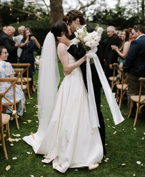 A bride and groom share a kiss at their outdoor wedding ceremony. The bride is wearing a white gown and holding a bouquet of white flowers, while the groom is in a black suit. Guests seated in wooden chairs and standing in the background are clapping and smiling.