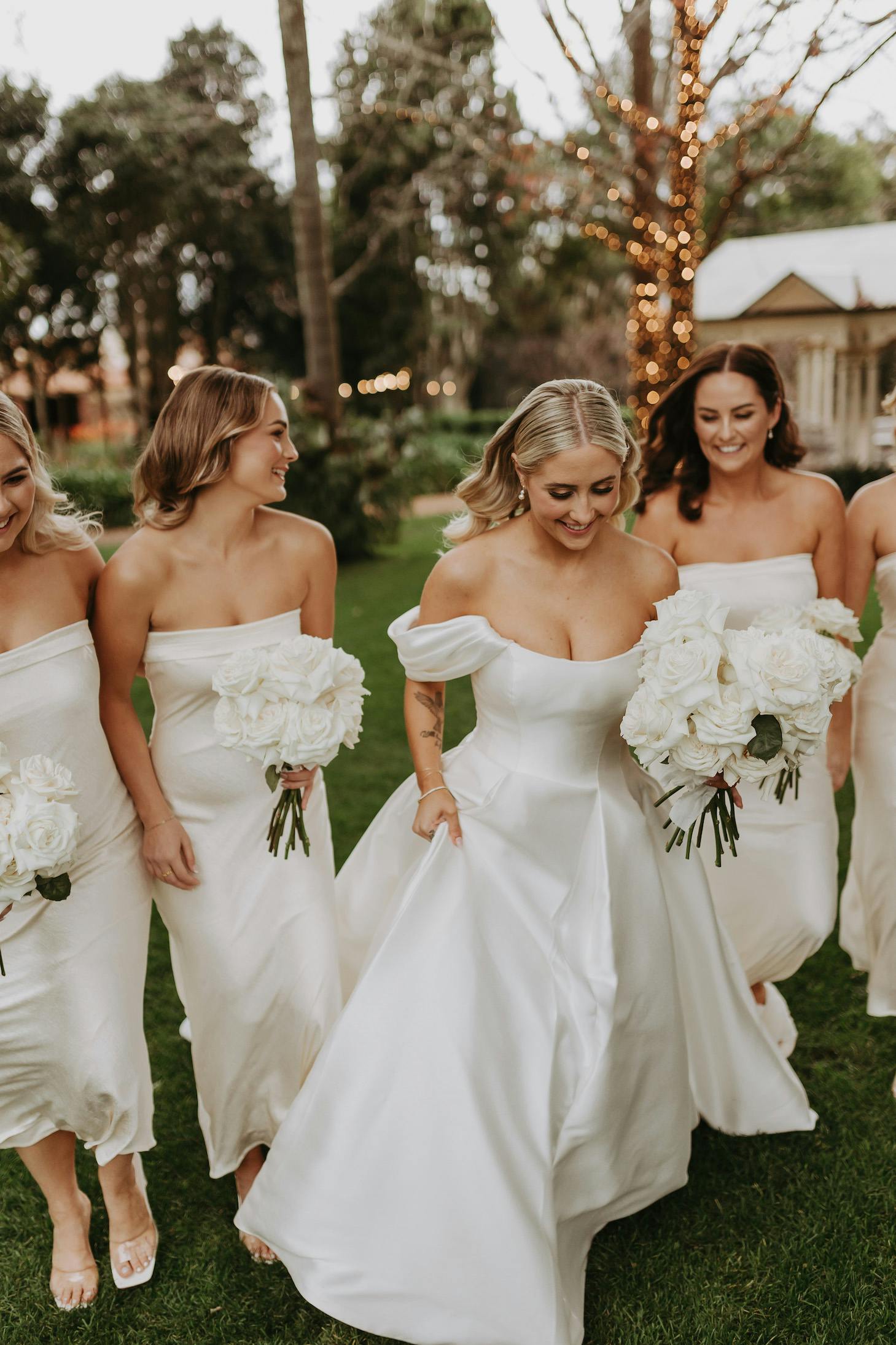 A bride in a white off-the-shoulder wedding dress walks on grass with her bridesmaids, who are also wearing white off-the-shoulder dresses. All are holding white rose bouquets. The background features trees and soft lighting.