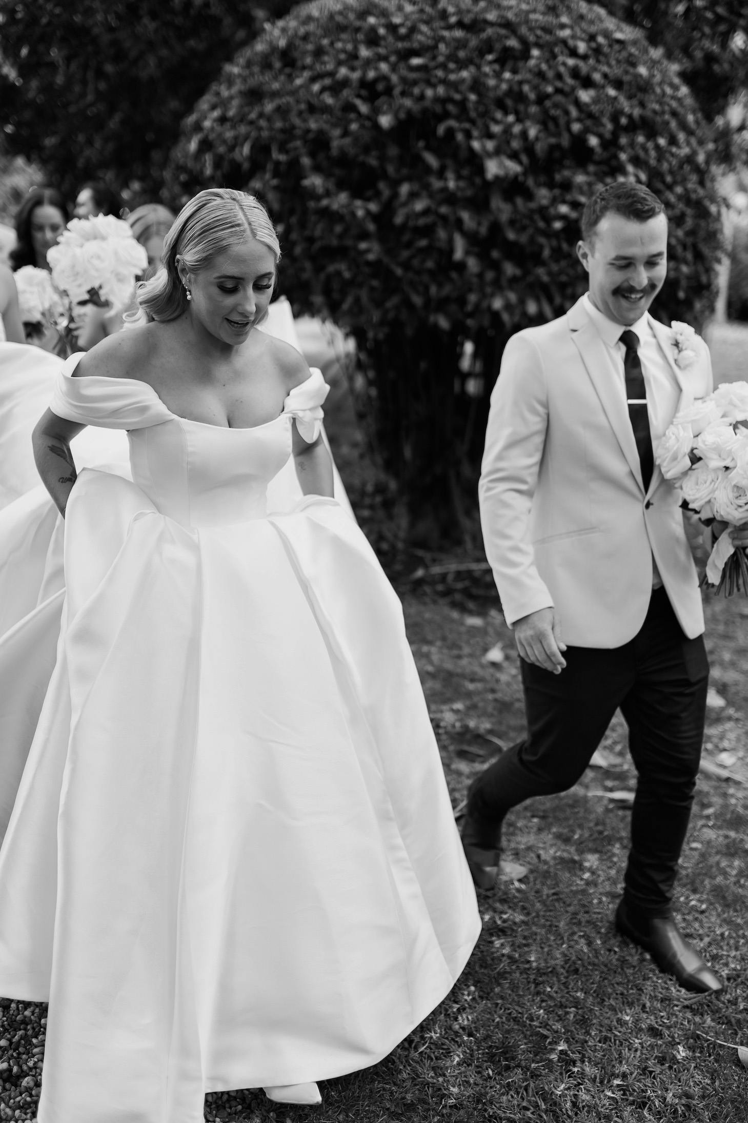 A black and white photo of a wedding couple walking outdoors. The bride wears an off-the-shoulder ball gown and smiles while holding her dress. The groom, dressed in a light suit jacket and dark pants, walks beside her carrying a bouquet. Bridesmaids follow in the background.