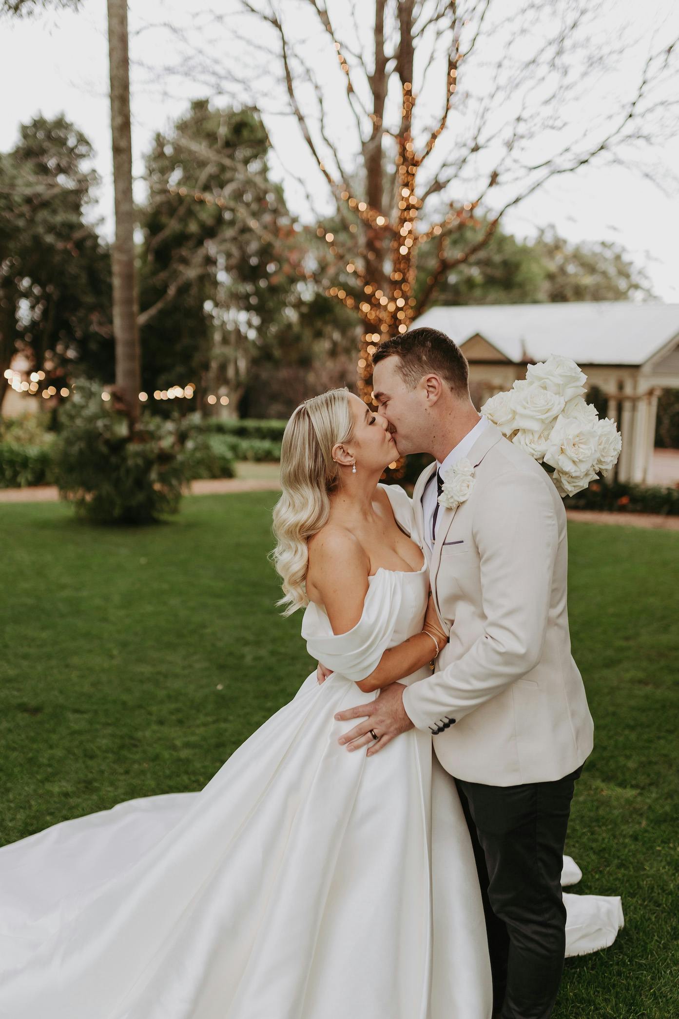 A bride and groom share a kiss on a lawn in front of a tree wrapped with lights. The bride is wearing an off-shoulder white wedding dress and holding a bouquet of white flowers, while the groom is dressed in a light-colored suit.