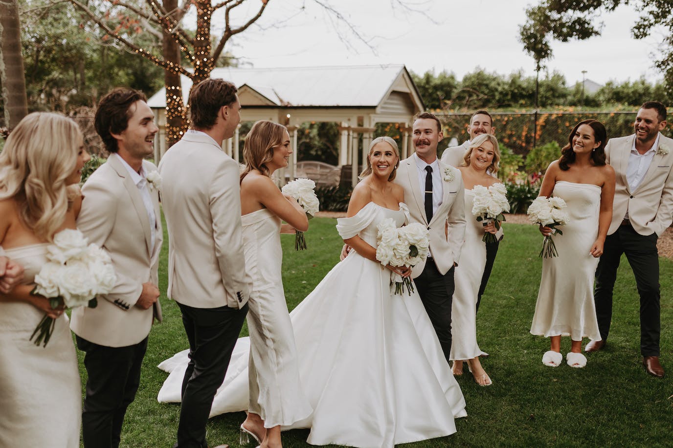 A joyful wedding party stands on a green lawn. The bride in a white gown holds a bouquet, surrounded by bridesmaids in white dresses and groomsmen in light-colored suits. Everyone is smiling, and the background features trees, a small building, and lit string lights.