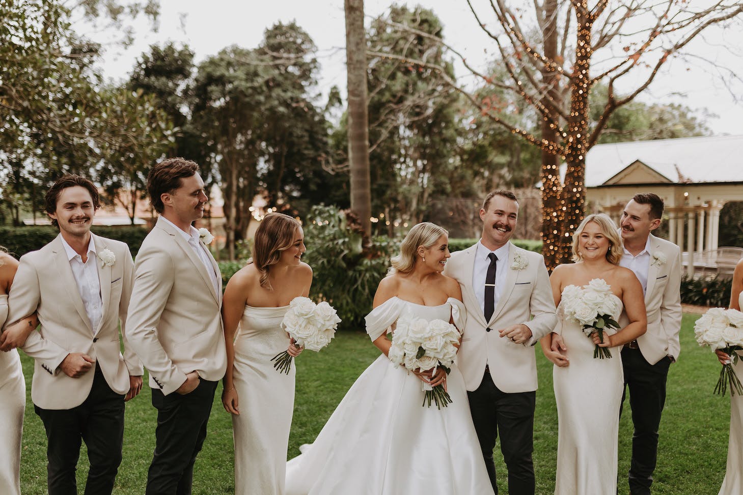 Seven people stand outdoors in formal attire next to one another, smiling and laughing. The group includes three men in light-colored suits with boutonnieres and four women in white dresses holding bouquets. They are in a garden setting with trees and a decorated tree trunk.