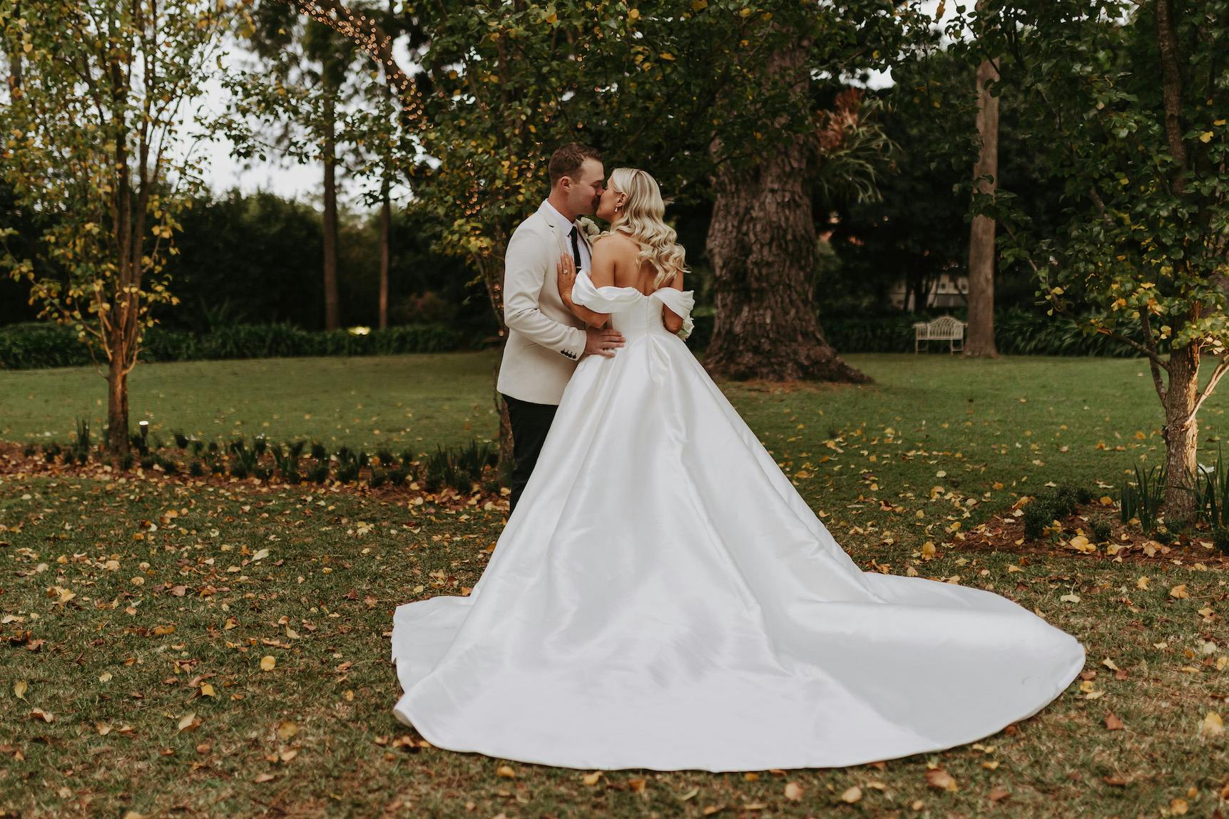 A bride and groom share a kiss outdoors in a lush garden. The bride wears a flowing white gown with a long train, and the groom is dressed in a white dinner jacket and black pants. They stand on grass with trees and foliage around them.