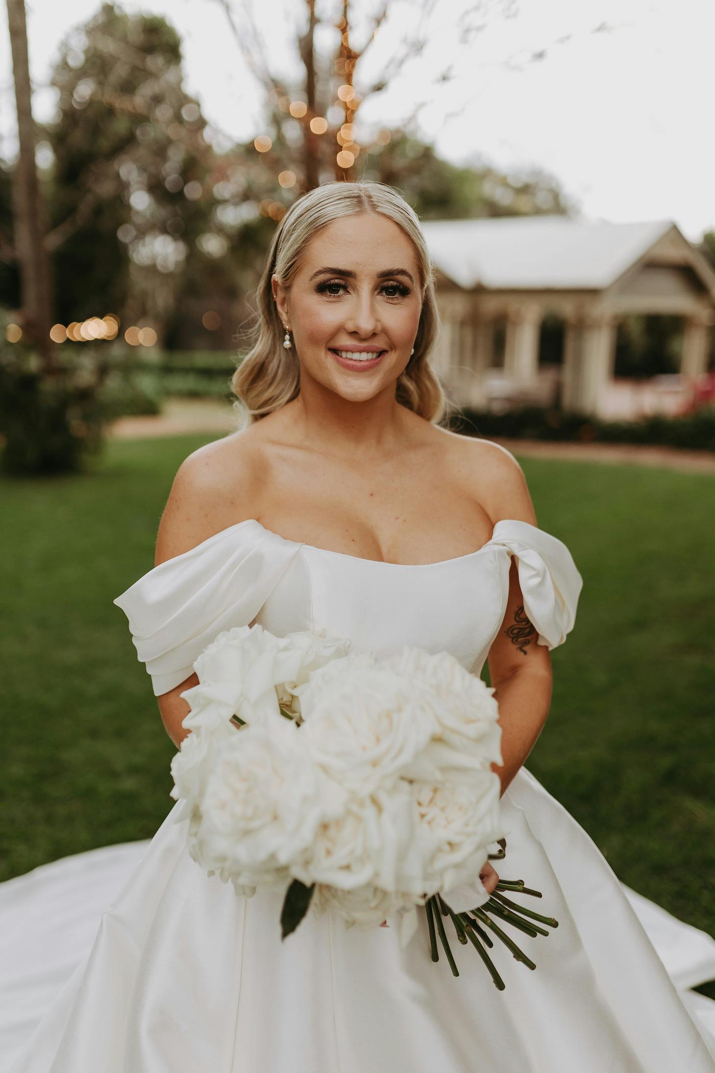 A bride with long blonde hair stands outside on a grassy lawn. She is wearing a white off-the-shoulder wedding dress and holding a bouquet of white roses. In the background, there are trees and a small building. The bride is smiling at the camera.