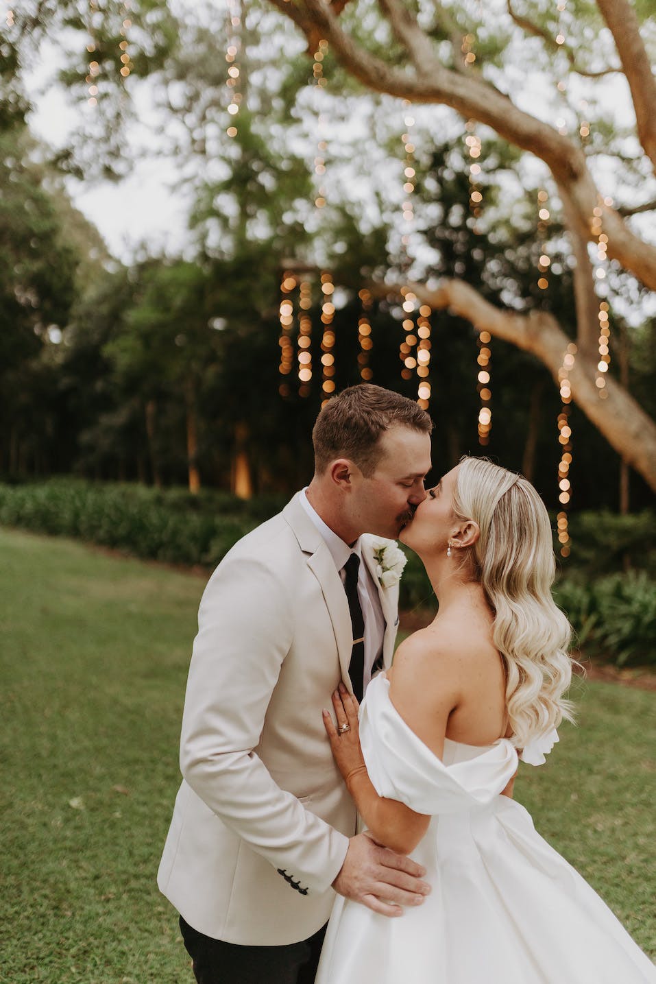 A couple in wedding attire share a kiss outdoors. The groom is wearing a beige suit, and the bride is in an off-the-shoulder white gown. They are standing in a grassy area with trees and string lights glowing in the background.