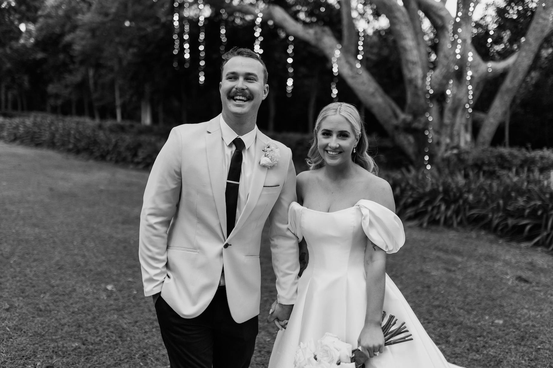 Black and white photo of a couple on their wedding day. The groom wears a light-colored suit and has a mustache, while the bride wears an off-shoulder wedding dress and holds a bouquet. They smile and stand outdoors with fairy lights hanging from trees in the background.