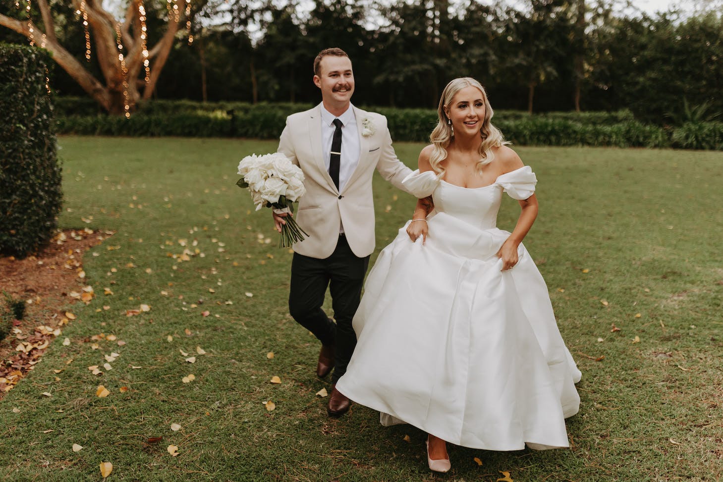 A smiling bride and groom walk hand in hand on a grassy lawn. The bride wears a white off-the-shoulder gown and holds her dress up in one hand while holding a bouquet of white flowers in the other. The groom dons a light-colored blazer, dark pants, and a black tie.