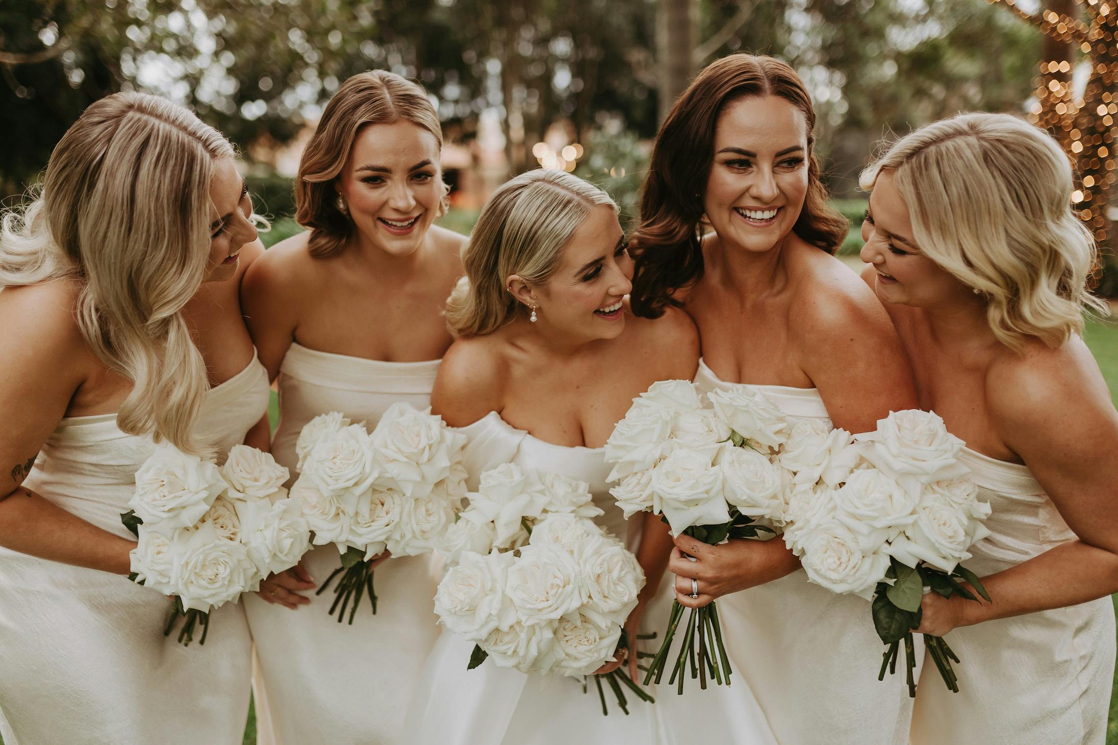 Five women in off-shoulder white dresses stand closely together, smiling and holding white rose bouquets. They appear joyful, with greenery and twinkling lights in the background, suggesting a festive or celebratory occasion.