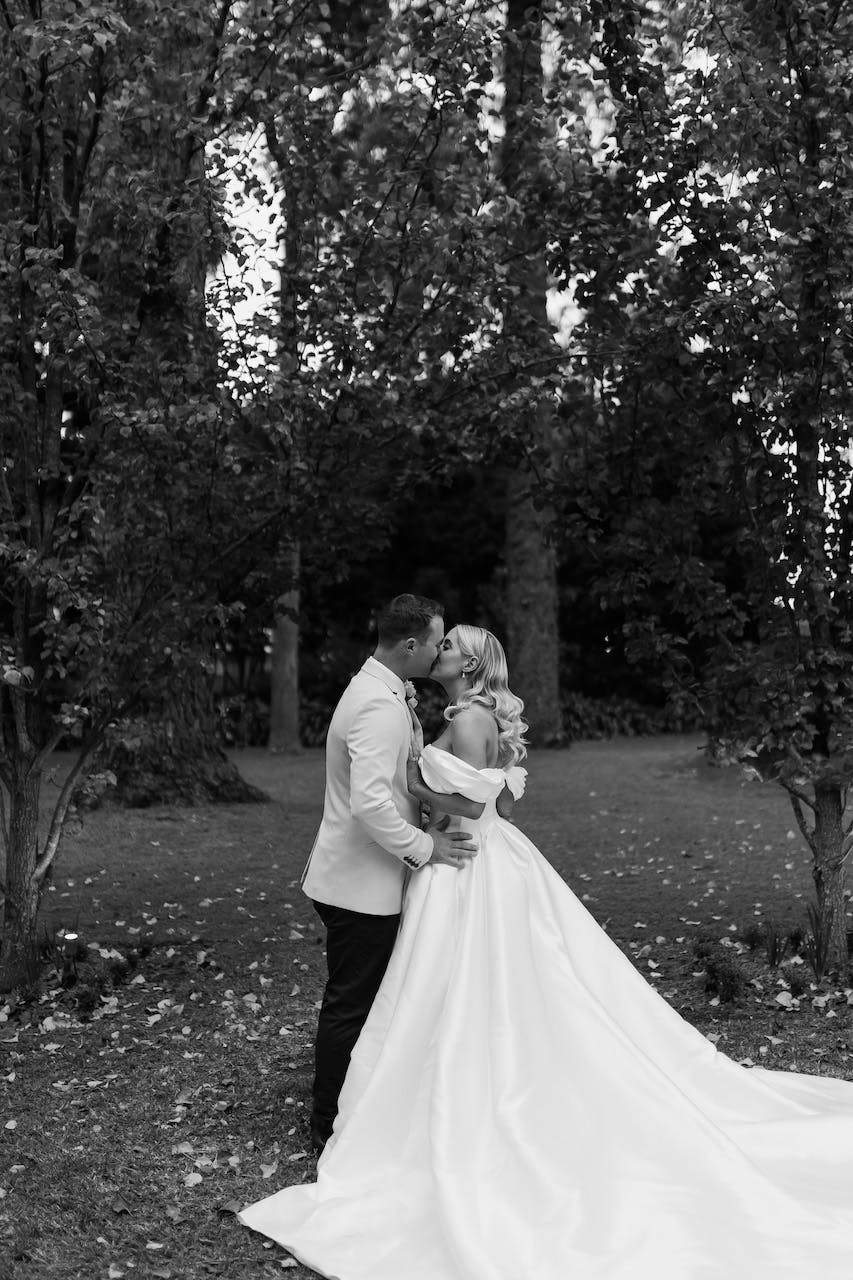 A bride and groom share a kiss outside in a forested area. The bride is wearing an off-shoulder wedding gown with a long train, while the groom is in a light-colored suit. They are surrounded by trees, and the ground is scattered with leaves. The image is in black and white.