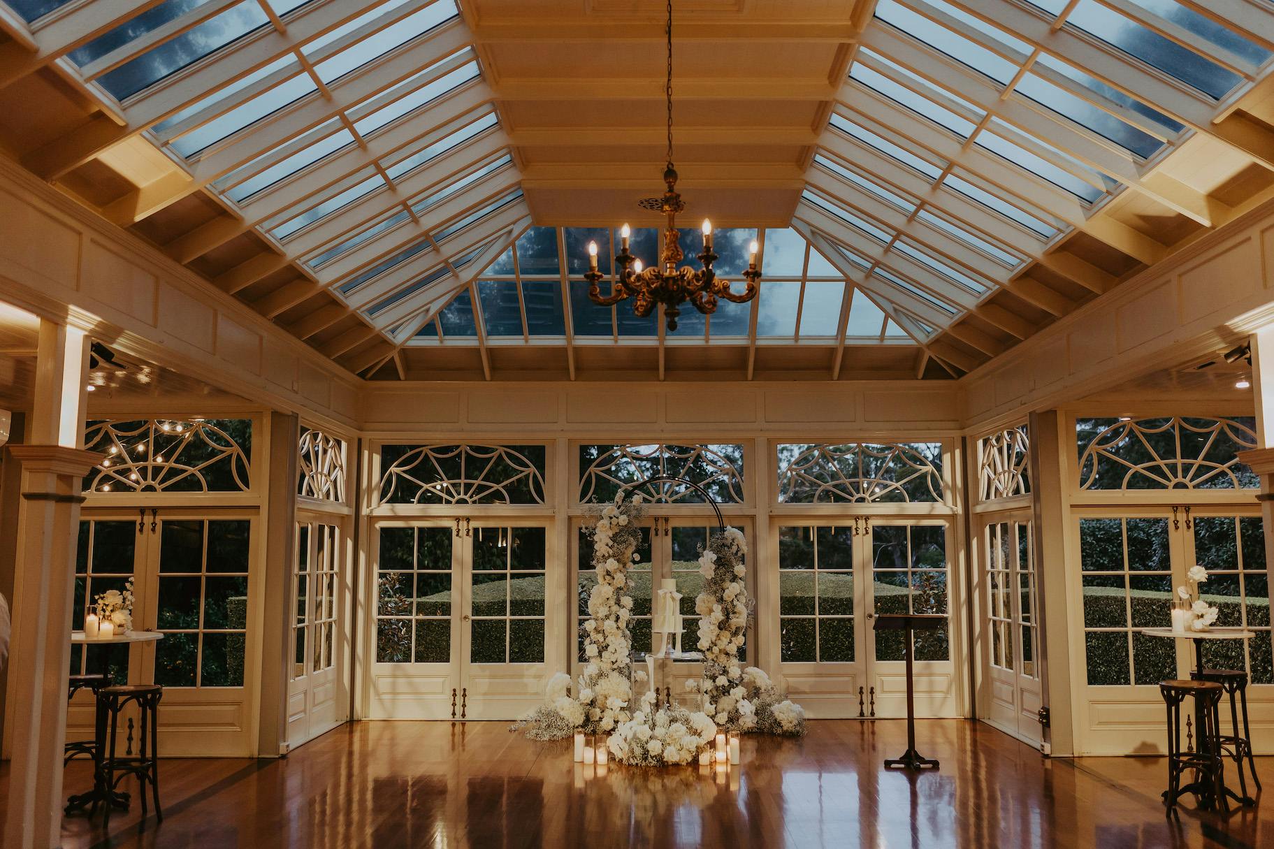 A spacious, elegant room with a glass ceiling and chandelier. Large windows and French doors create a bright atmosphere. White floral arrangements with candles surround a central display on a polished wood floor, ready for a formal event or ceremony.