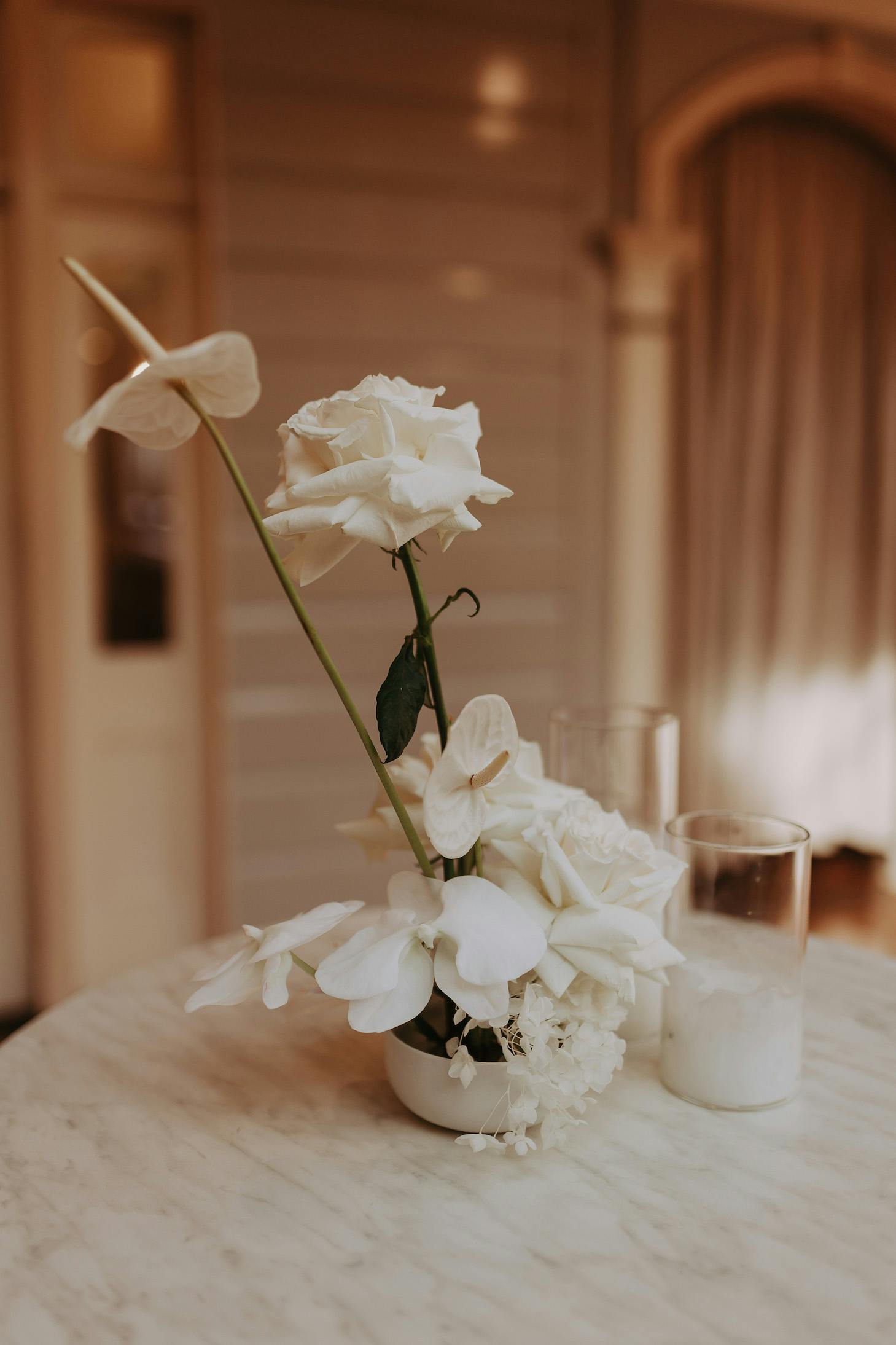 A small floral arrangement sits on a marble table, featuring white roses, white anthuriums, and other white flowers. Two glass candle holders, each containing a white candle, are positioned next to the floral display. The background shows part of an elegant room.